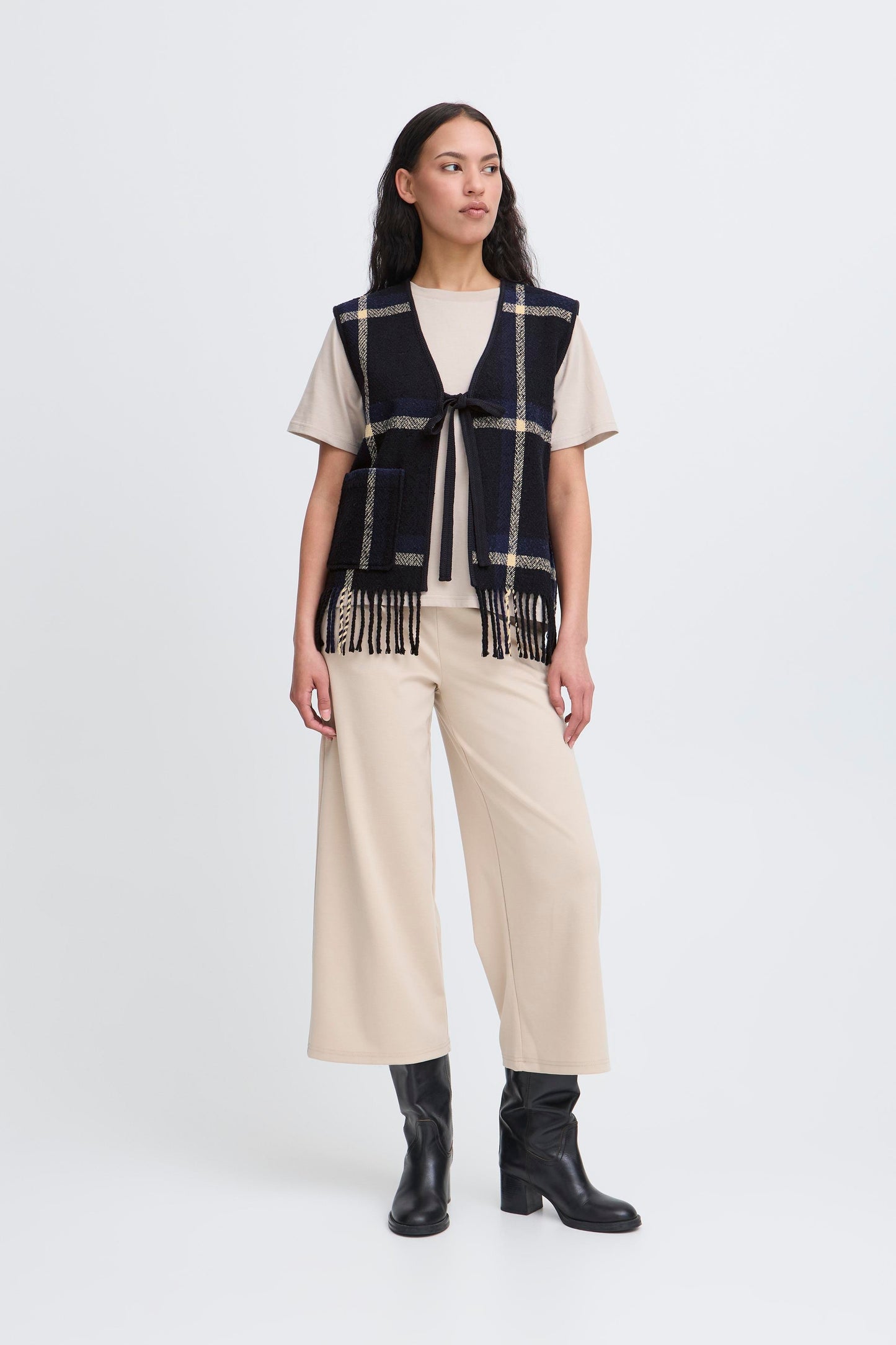 Woman wearing a plaid vest, white shirt, beige pants, and black boots on a white background