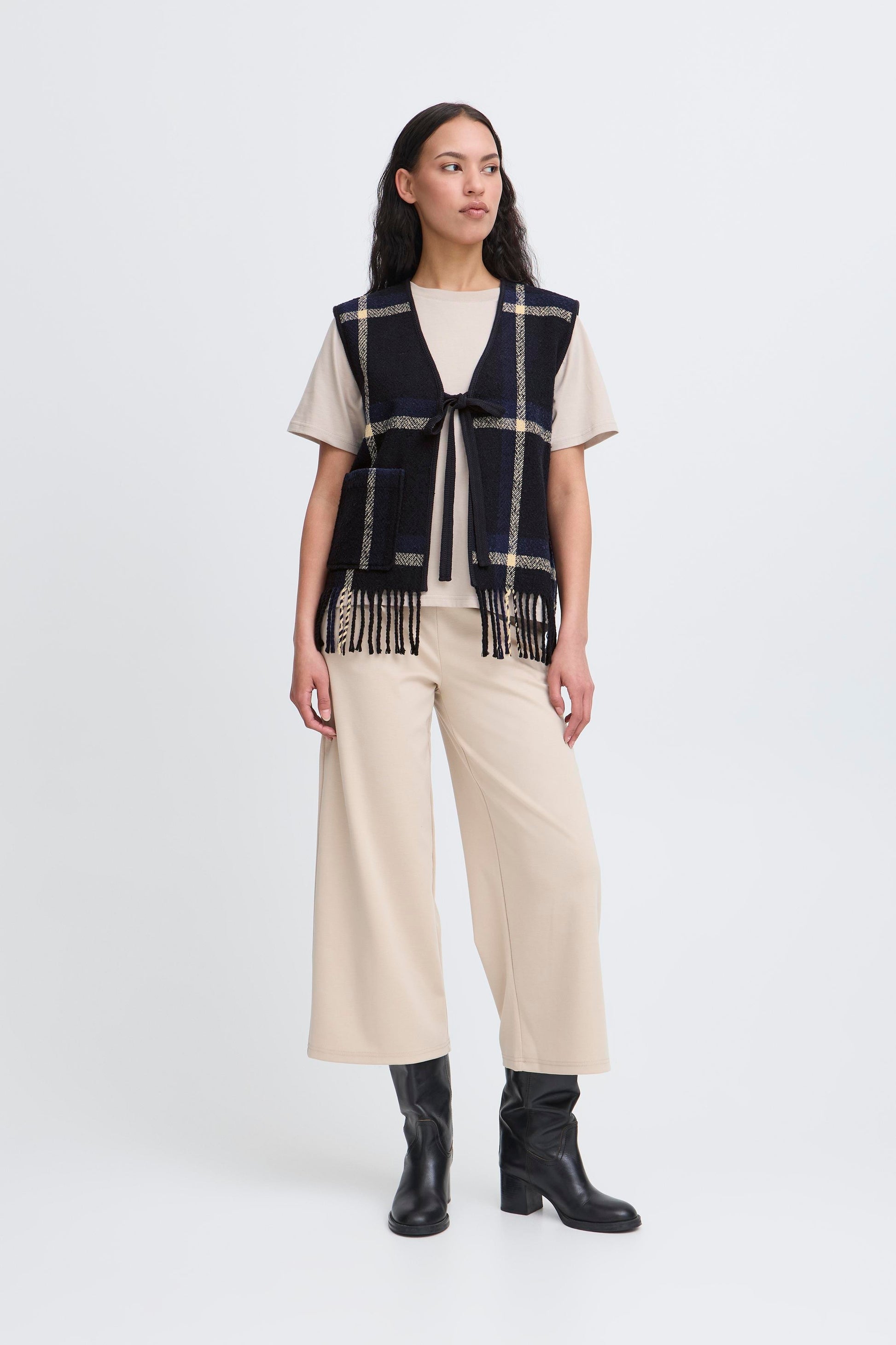 Woman wearing a plaid vest, white shirt, beige pants, and black boots on a white background
