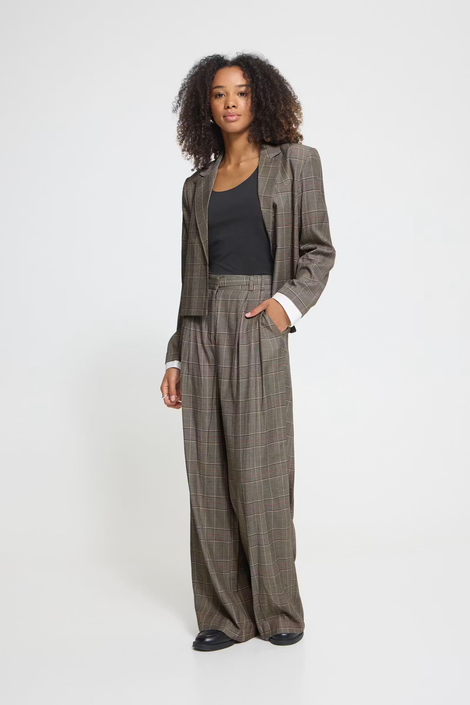 A woman stands against a light gray studio background wearing a tailored two-piece suit in a brown and gray plaid pattern, paired with a black top and black shoes.