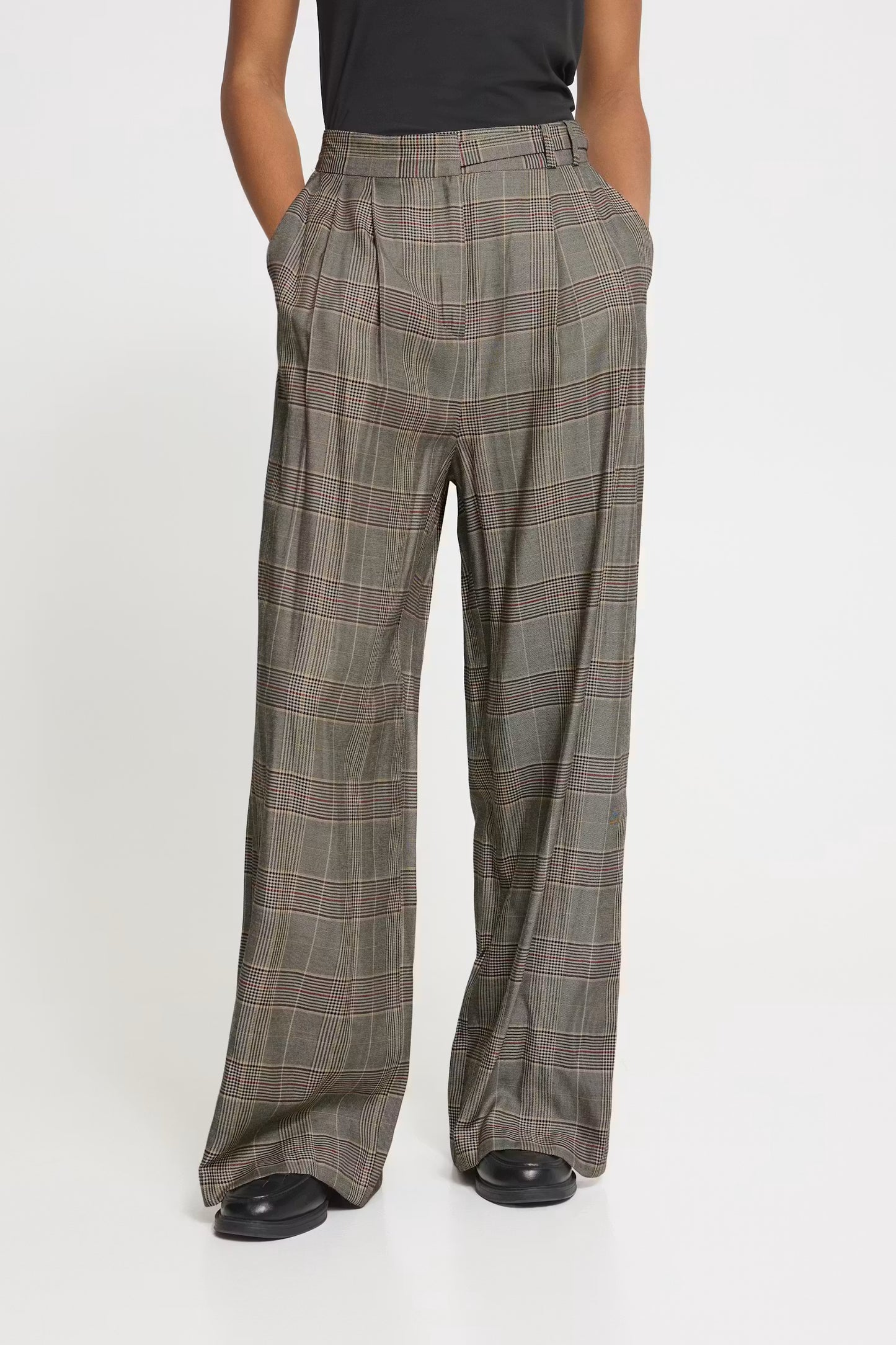 A person wearing high-waisted, wide-leg plaid trousers in a muted olive-gray color with thin red and beige windowpane lines, paired with a black top and black shoes, against a plain white studio background.