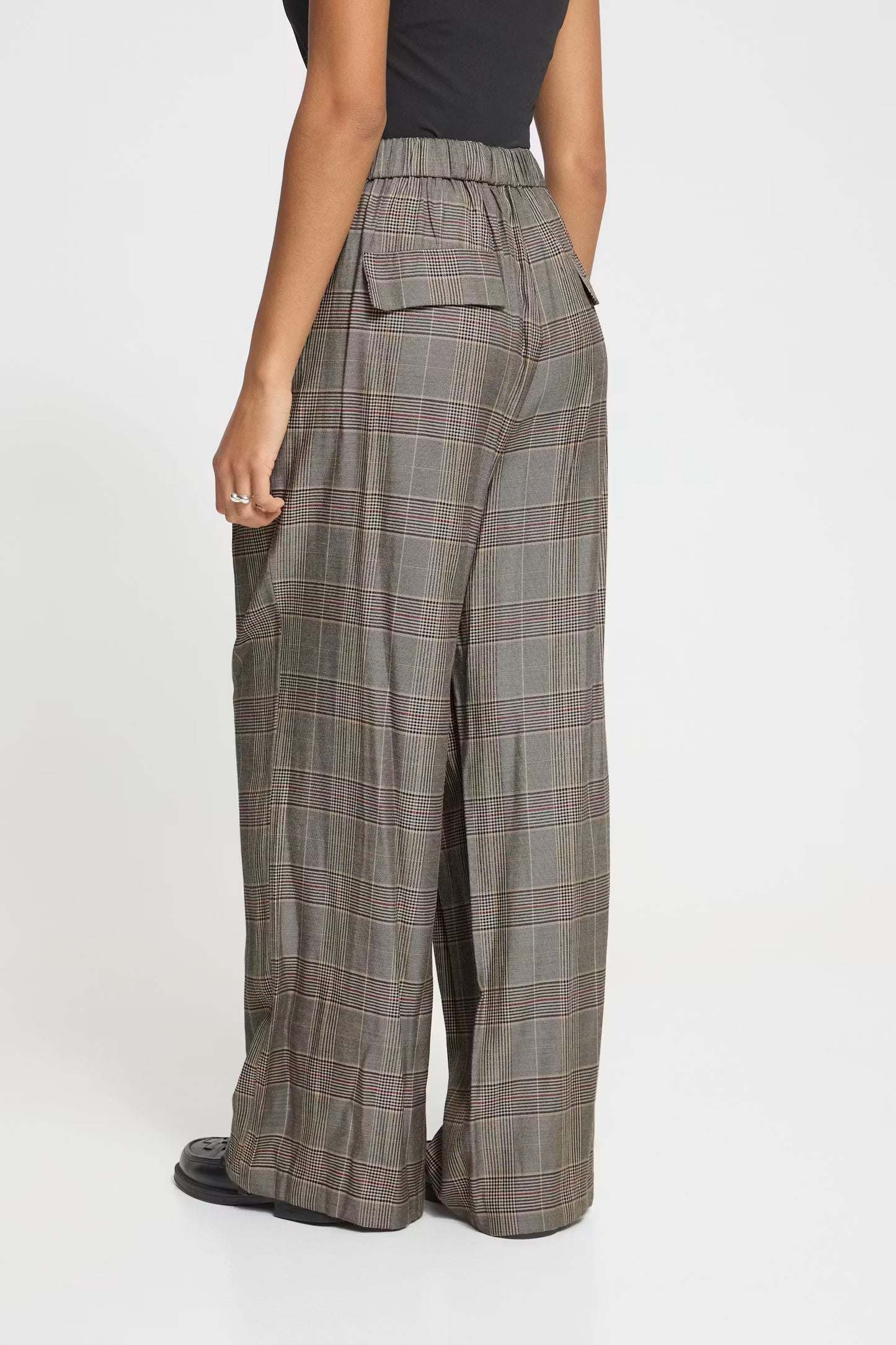 A model shown from the back wearing high-waisted, wide-leg plaid trousers in a gray and brown tartan pattern, paired with a black top and black shoes, against a plain white studio background.