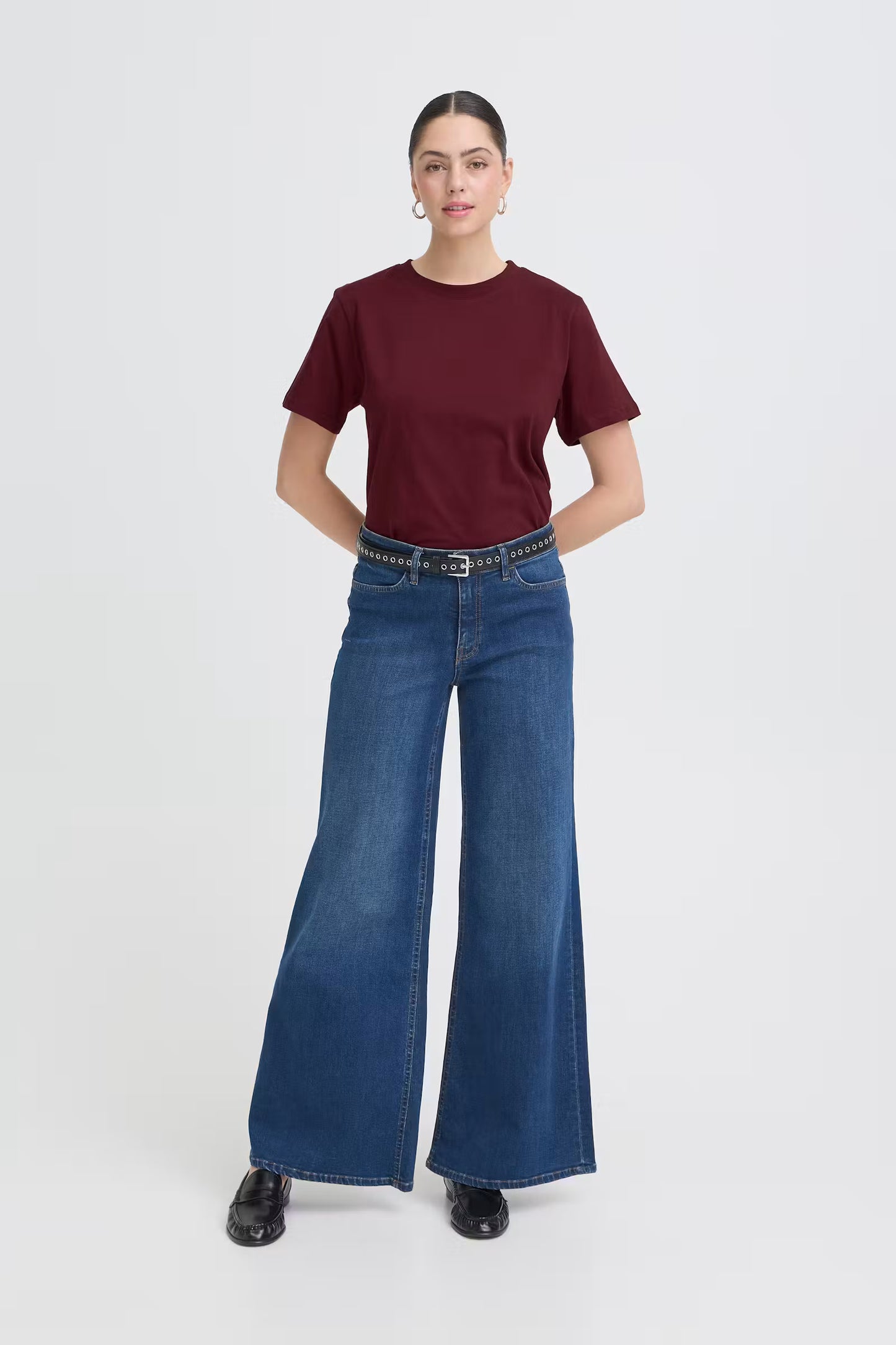 A woman modeling a short-sleeve crewneck t-shirt in burgundy and wide-leg blue jeans with a black belt, standing against a light gray studio background.