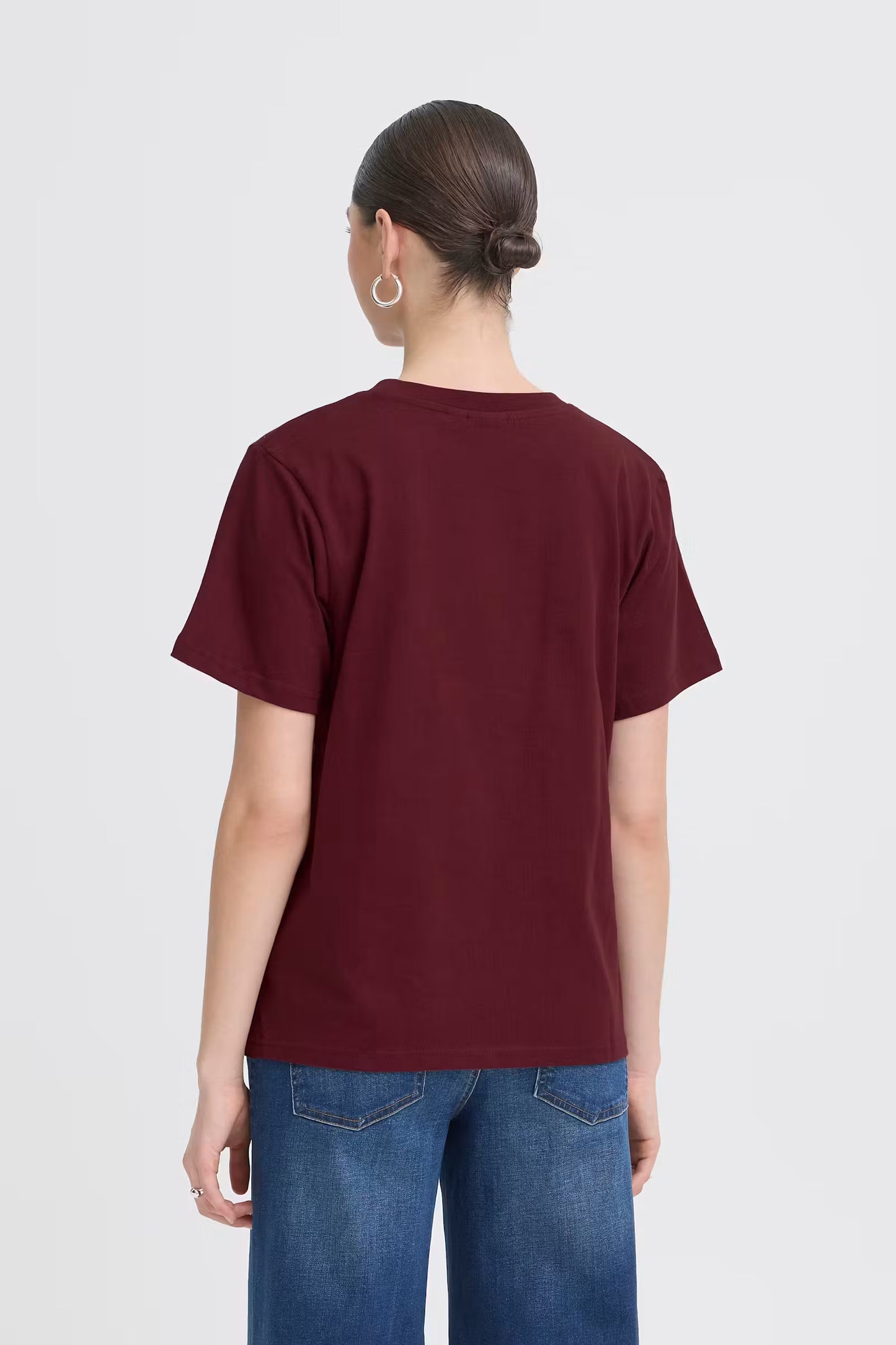 A woman viewed from the back wearing a short-sleeve crew-neck t-shirt in burgundy and blue jeans, set against a plain light gray studio background.