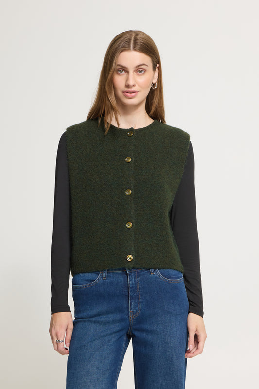 A woman modeling a dark green sleeveless button-front knit vest over a black long-sleeve top and blue jeans, photographed against a plain light gray studio background.