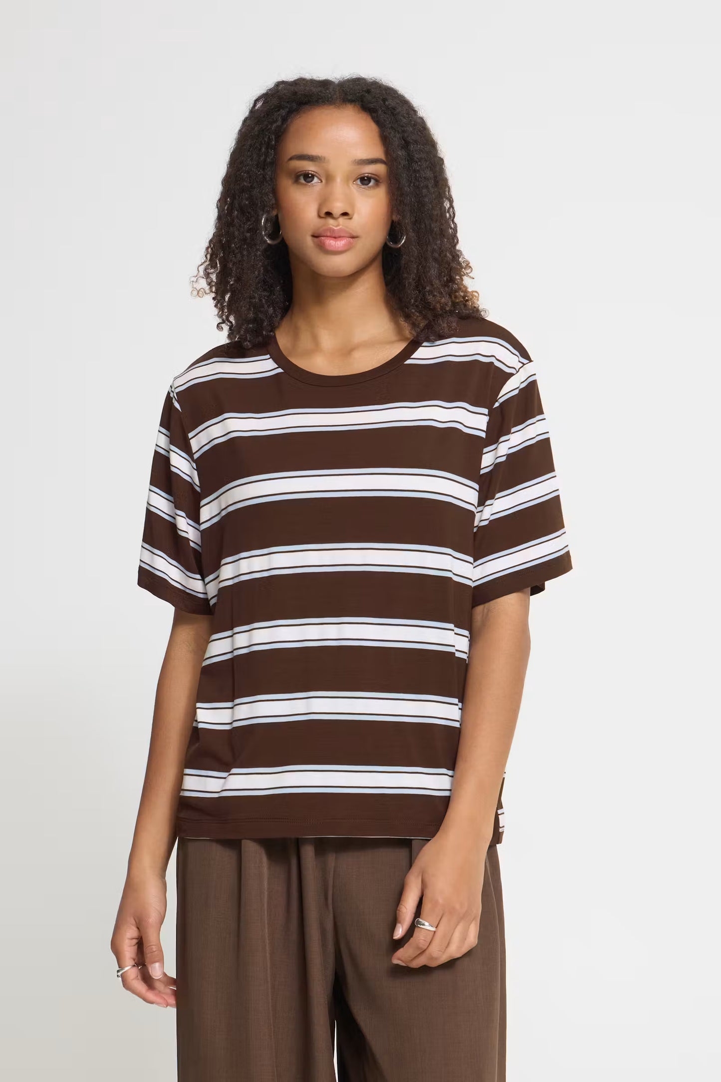 A woman modeling a brown and white horizontally striped short-sleeve t-shirt with brown pants, standing against a light gray studio background.