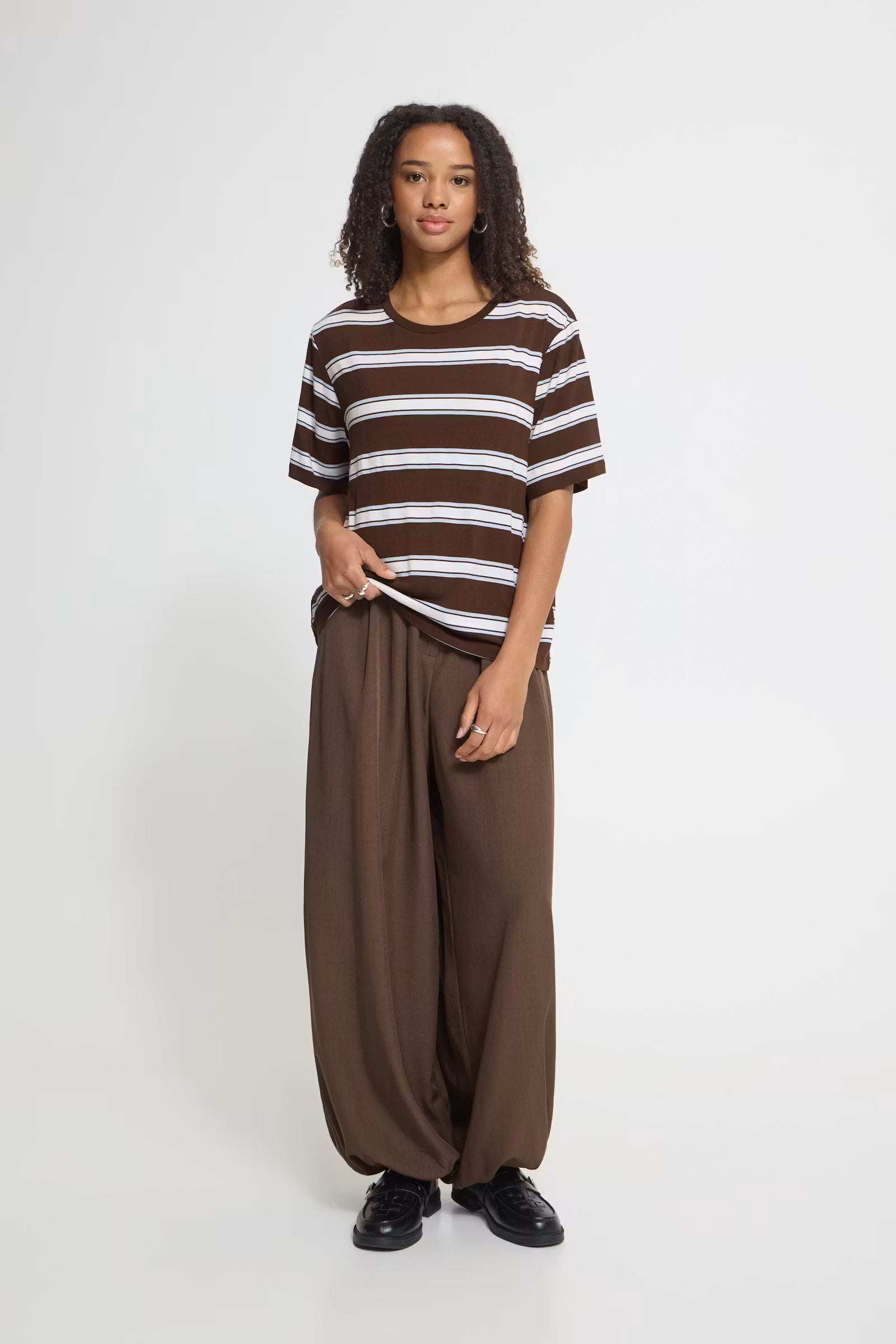 A woman modeling a brown and white horizontally striped short-sleeve t-shirt paired with loose-fitting brown wide-leg pants and black shoes, standing against a plain light gray studio background.