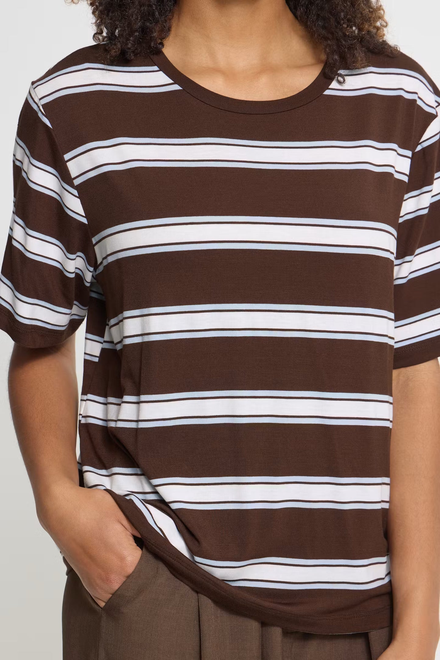 A close-up of a person wearing a brown short-sleeve crewneck t-shirt with horizontal white and thin blue stripes, paired with brown pants.