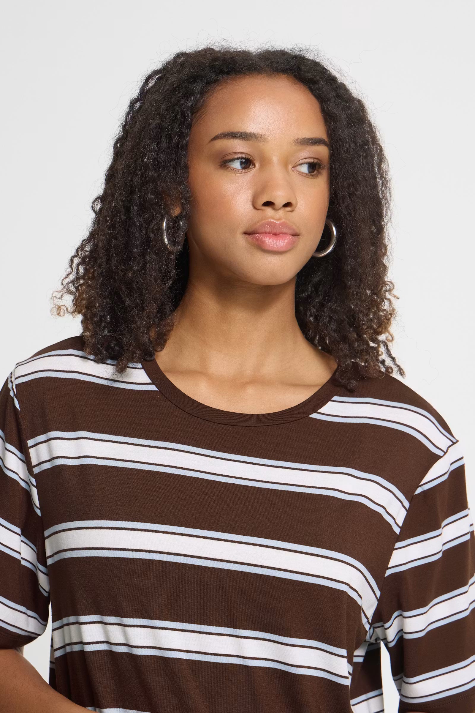 A woman with curly dark hair wearing a brown short-sleeve crewneck t-shirt with horizontal white and thin black stripes, photographed against a light gray studio background.