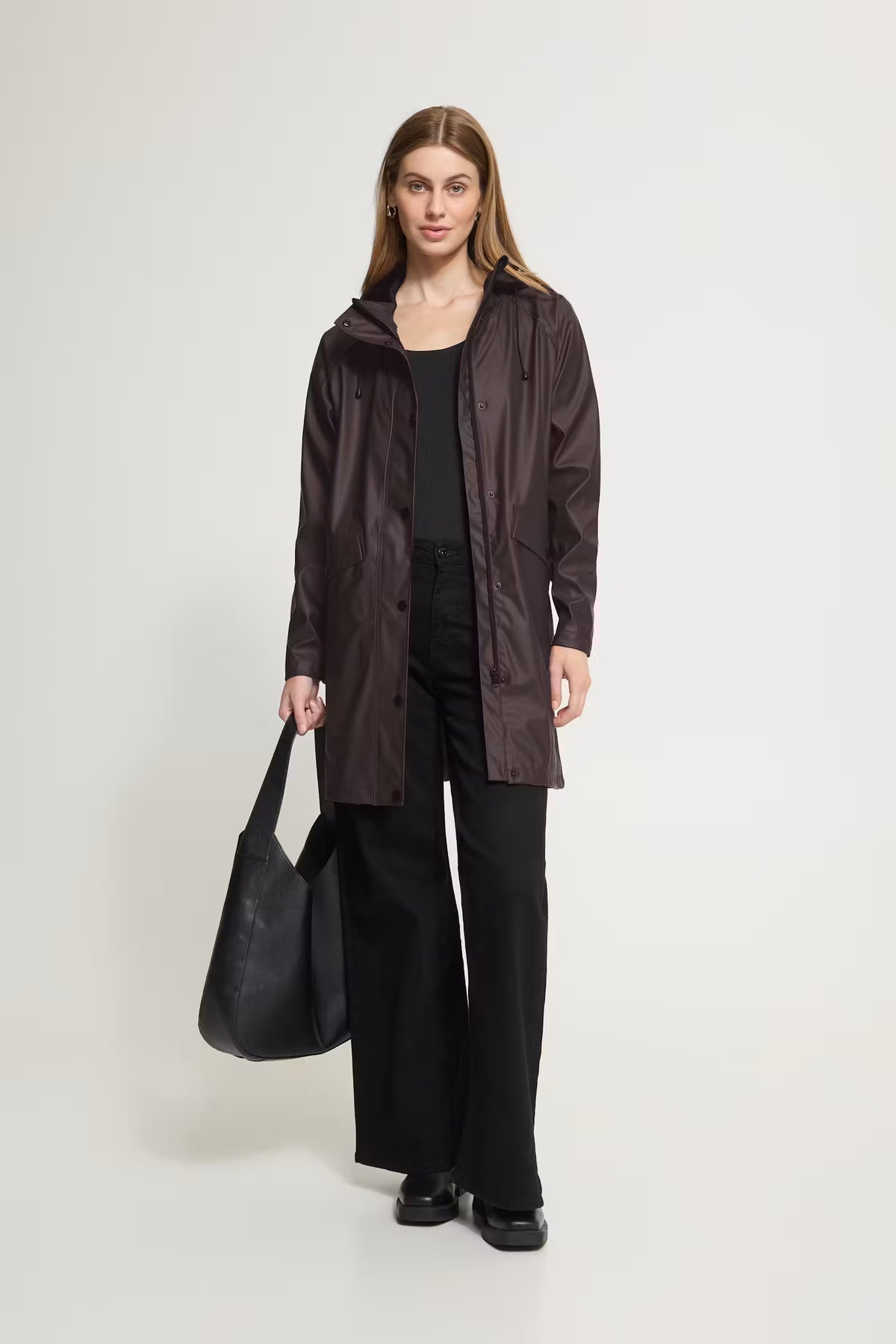 A woman stands against a light gray studio background wearing a dark brown hooded jacket over a black top and black wide-leg pants, while holding a large black tote bag.