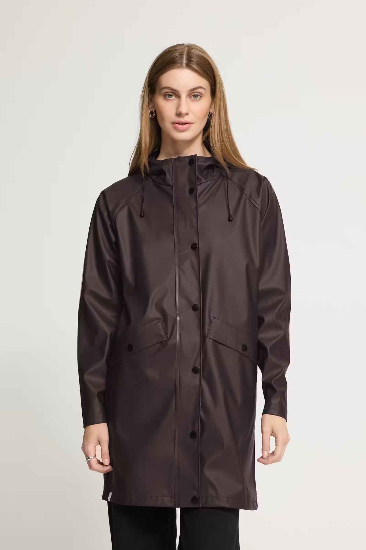 A woman modeling a dark brown hooded raincoat with a button-up front and front pockets, photographed against a plain light gray studio background.