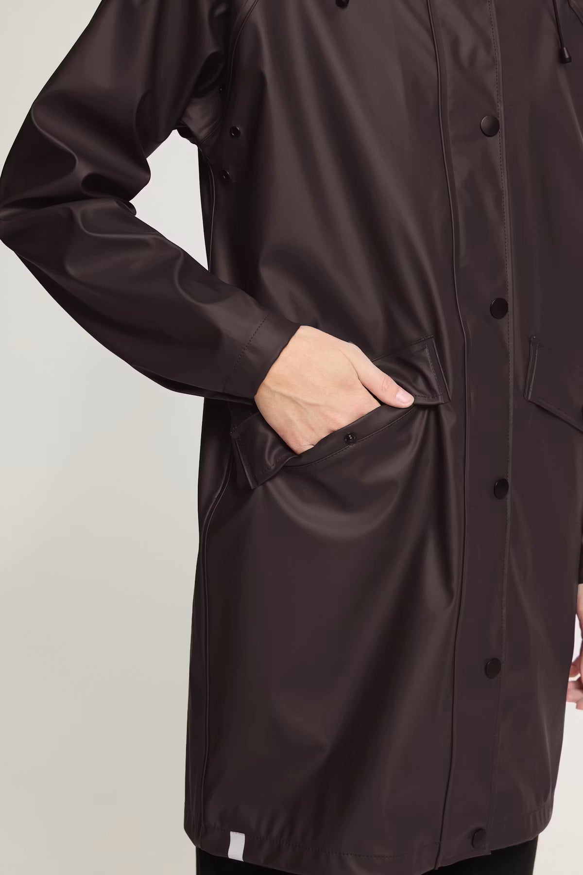 A close-up of a person wearing a dark brown waterproof raincoat with a matte finish, featuring a button-up front, side pockets, and a hand tucked into one of the pockets.