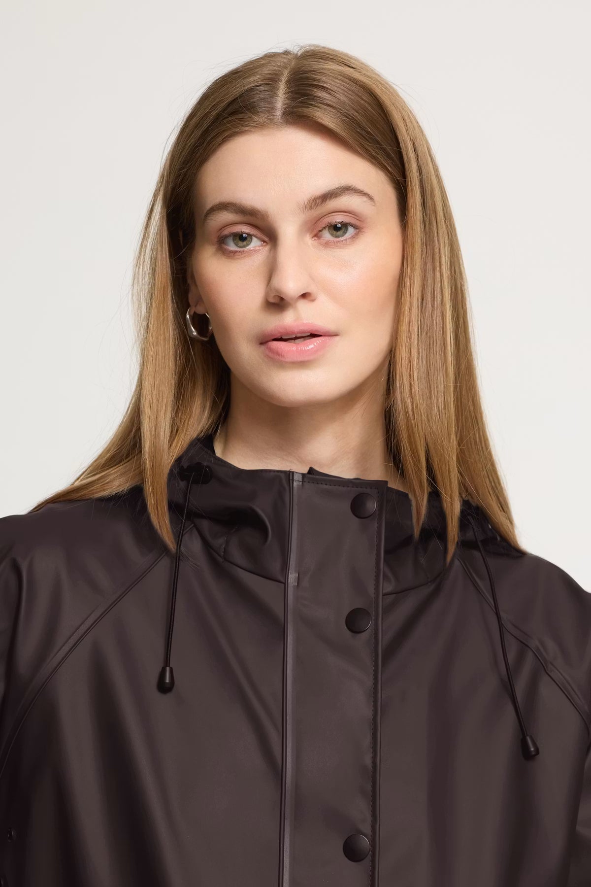 A woman with long light brown hair wearing a dark brown hooded rain jacket, shown from the chest up against a light gray studio background.