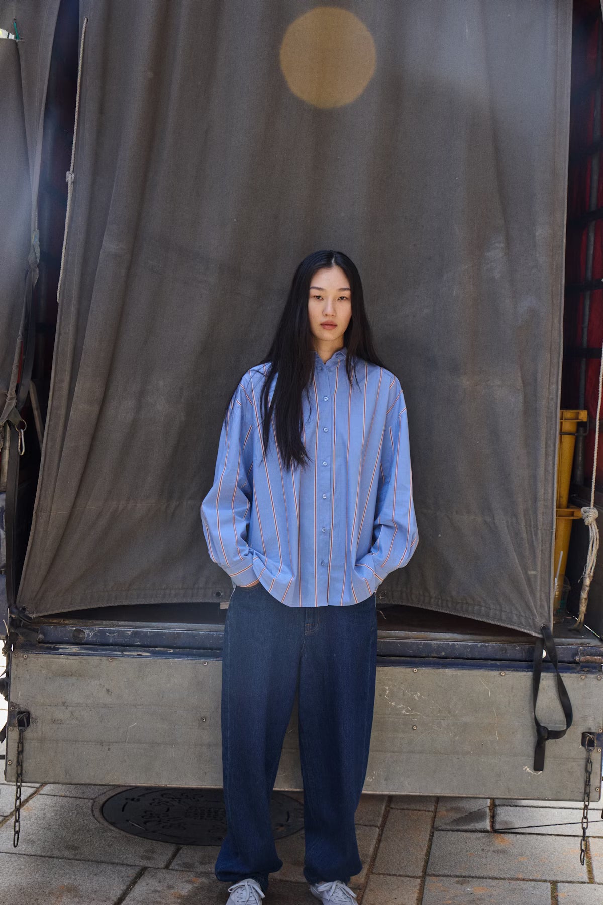 Person wearing a blue striped shirt and dark pants standing in front of a large gray curtain.