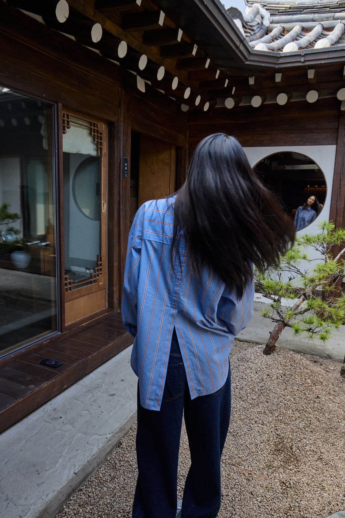 Person with long hair wearing a blue shirt standing in a traditional Japanese house courtyard.