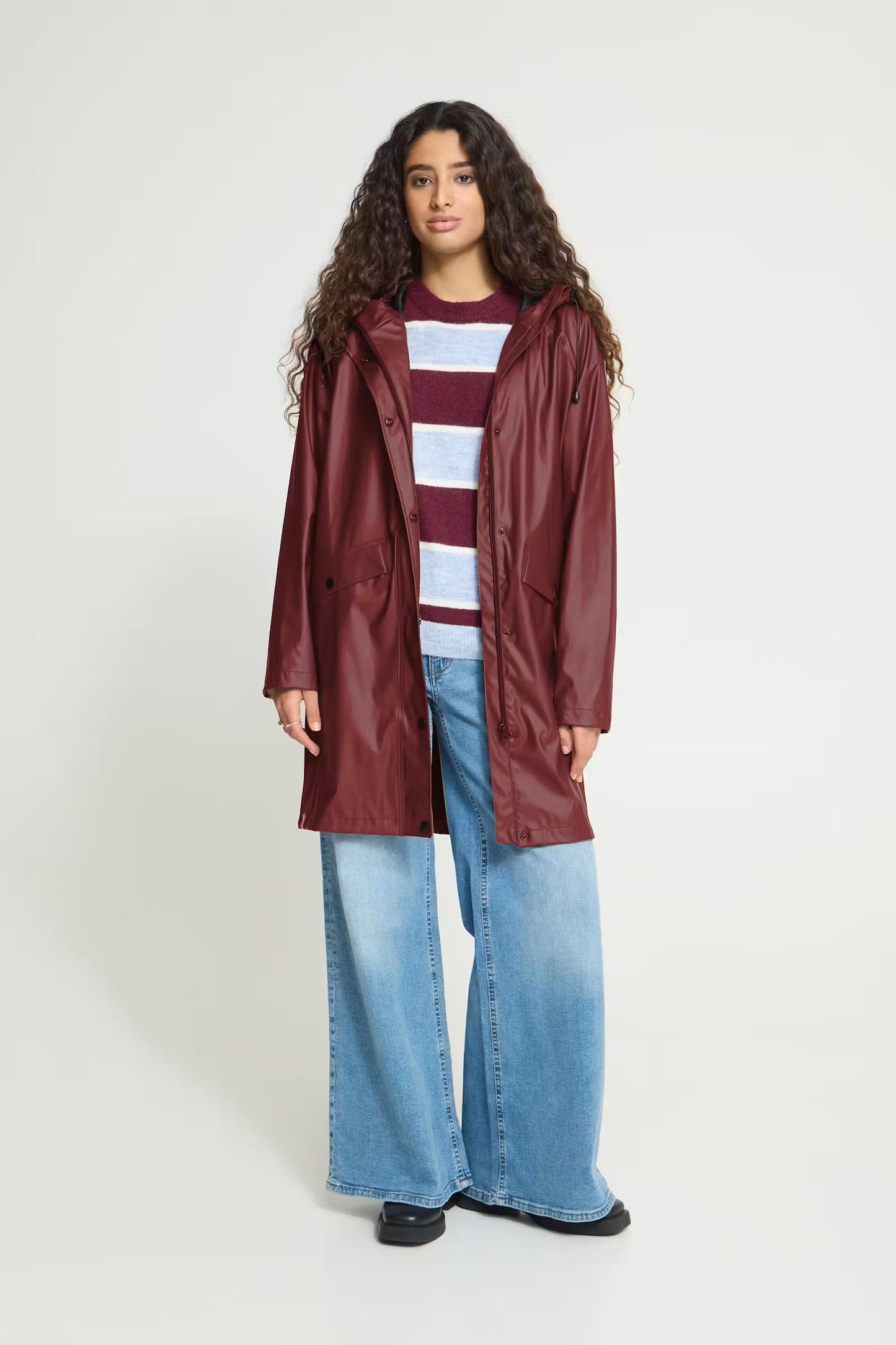 A woman with long curly brown hair wearing a glossy, long maroon raincoat and light blue jeans, standing against a light gray studio background.