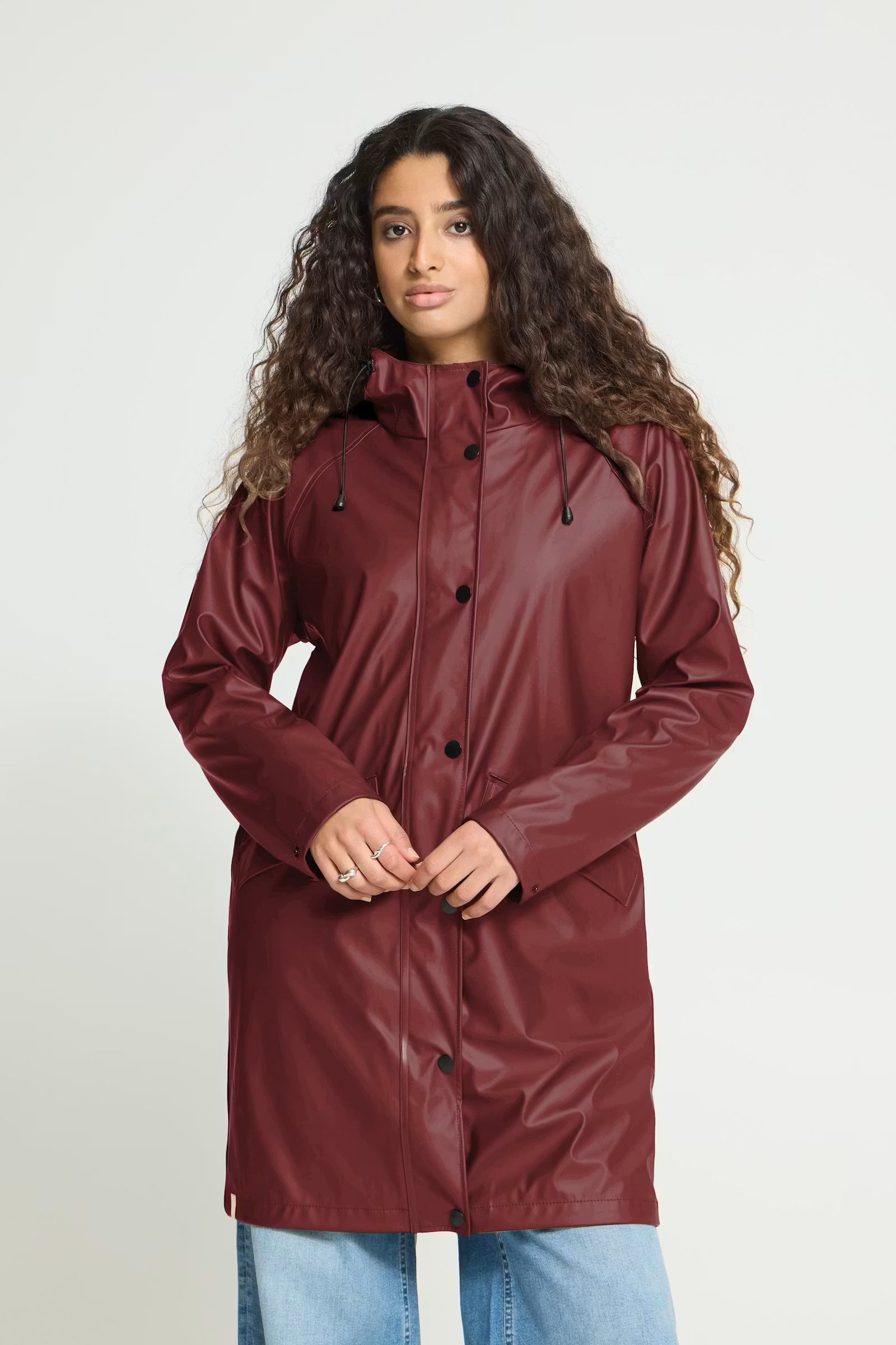A woman with long curly brown hair wearing a glossy, long maroon raincoat and light blue jeans, standing against a light gray studio background.
