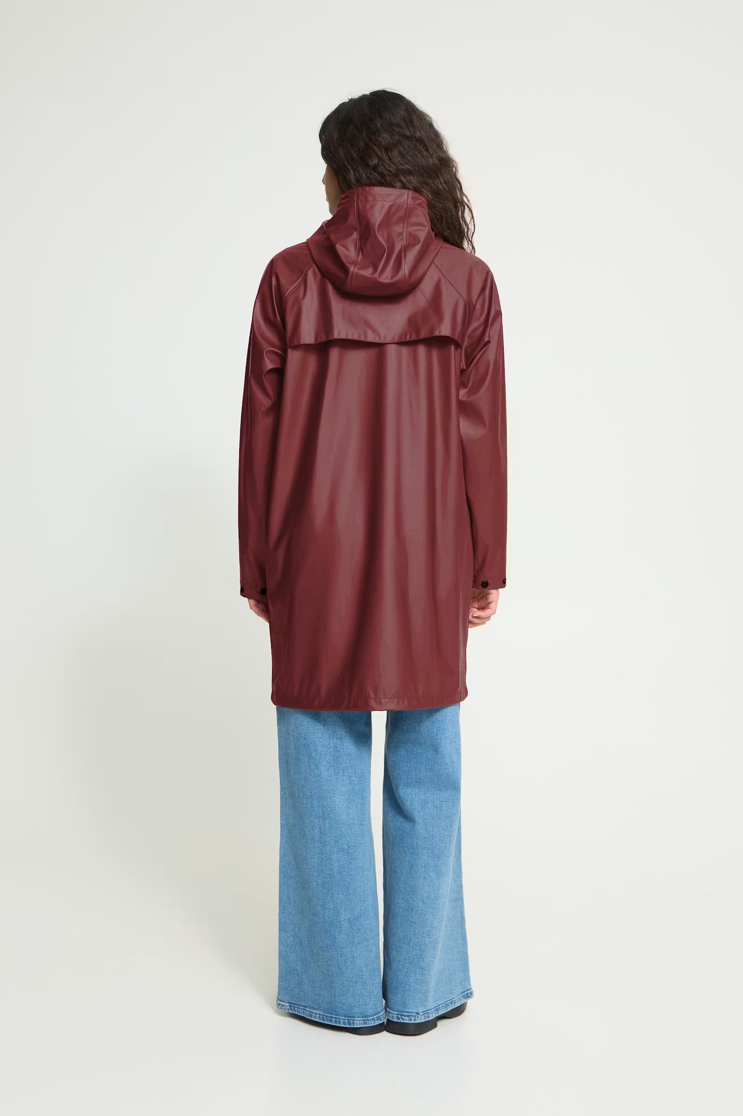 A model viewed from the back wearing a hooded raincoat in a deep burgundy color, paired with wide-leg blue jeans, against a plain white studio background.