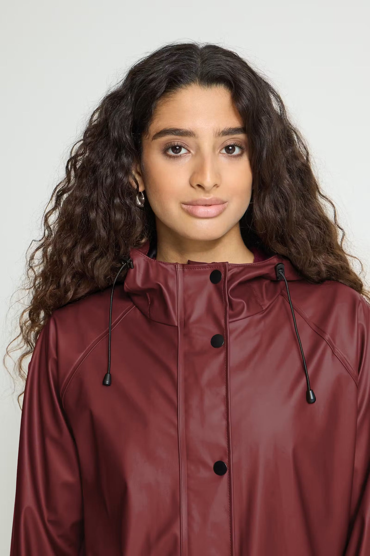 A woman with long, curly dark brown hair wearing a dark burgundy faux-leather hooded raincoat with black buttons and drawstrings, photographed against a light gray studio background.