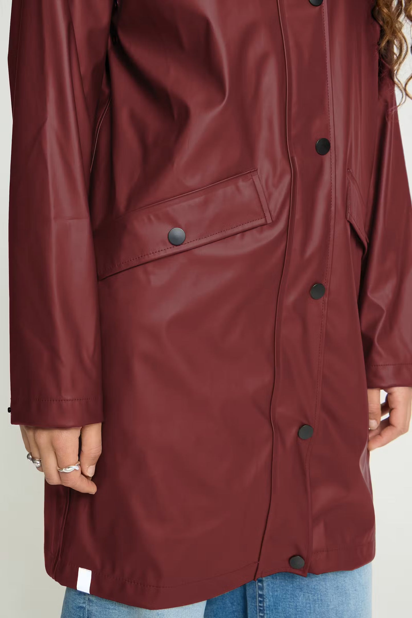 A close-up of a person wearing a glossy burgundy raincoat with dark buttons, paired with blue jeans, against a plain light background.