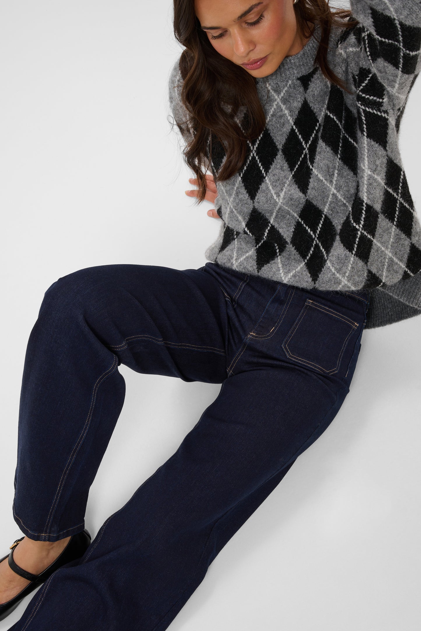 A woman wearing dark navy wide-leg jeans and a gray and black argyle sweater, posed against a light gray studio background.