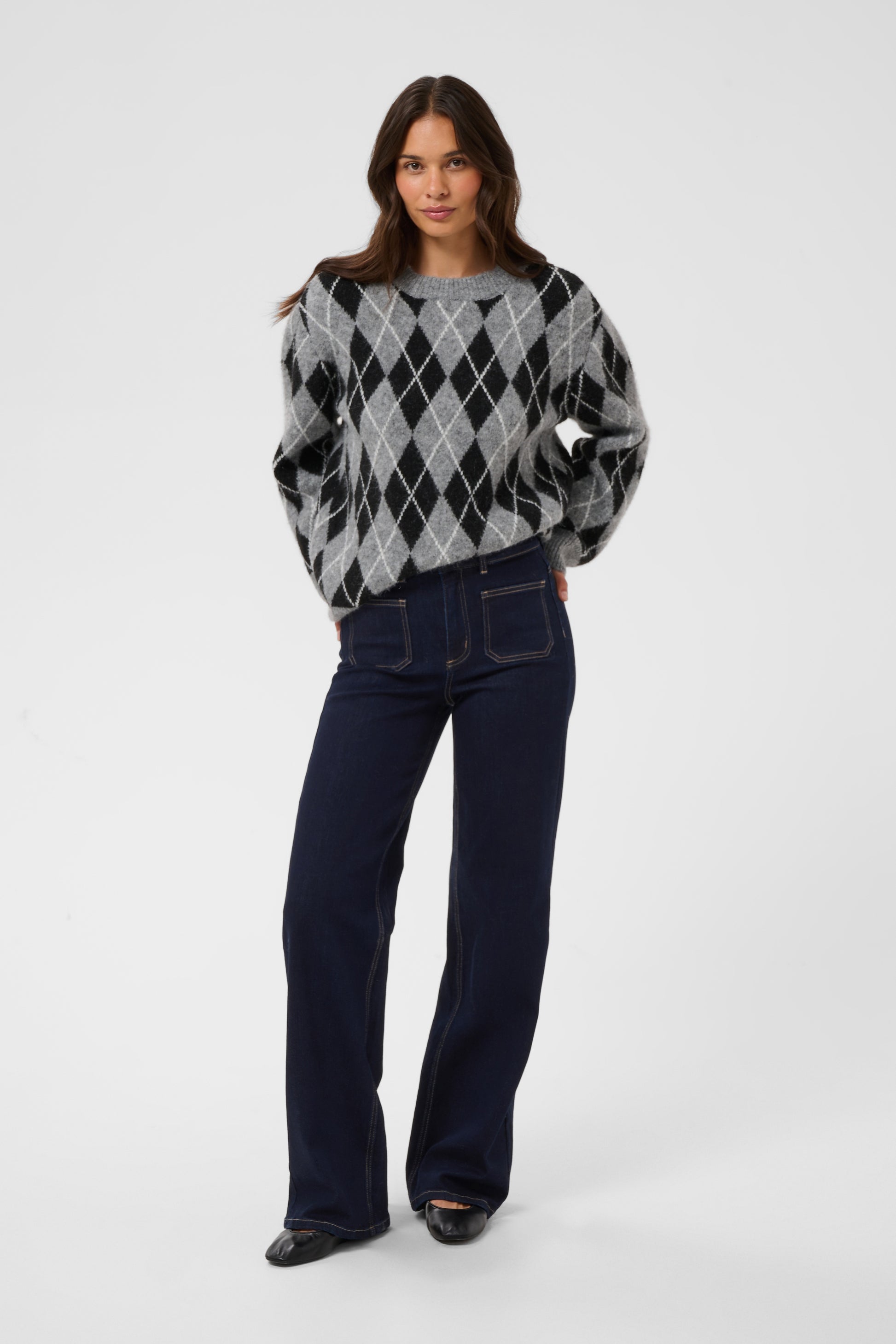 A woman modeling a gray and black argyle knit sweater with dark navy wide-leg jeans and black shoes, standing against a light gray studio background.