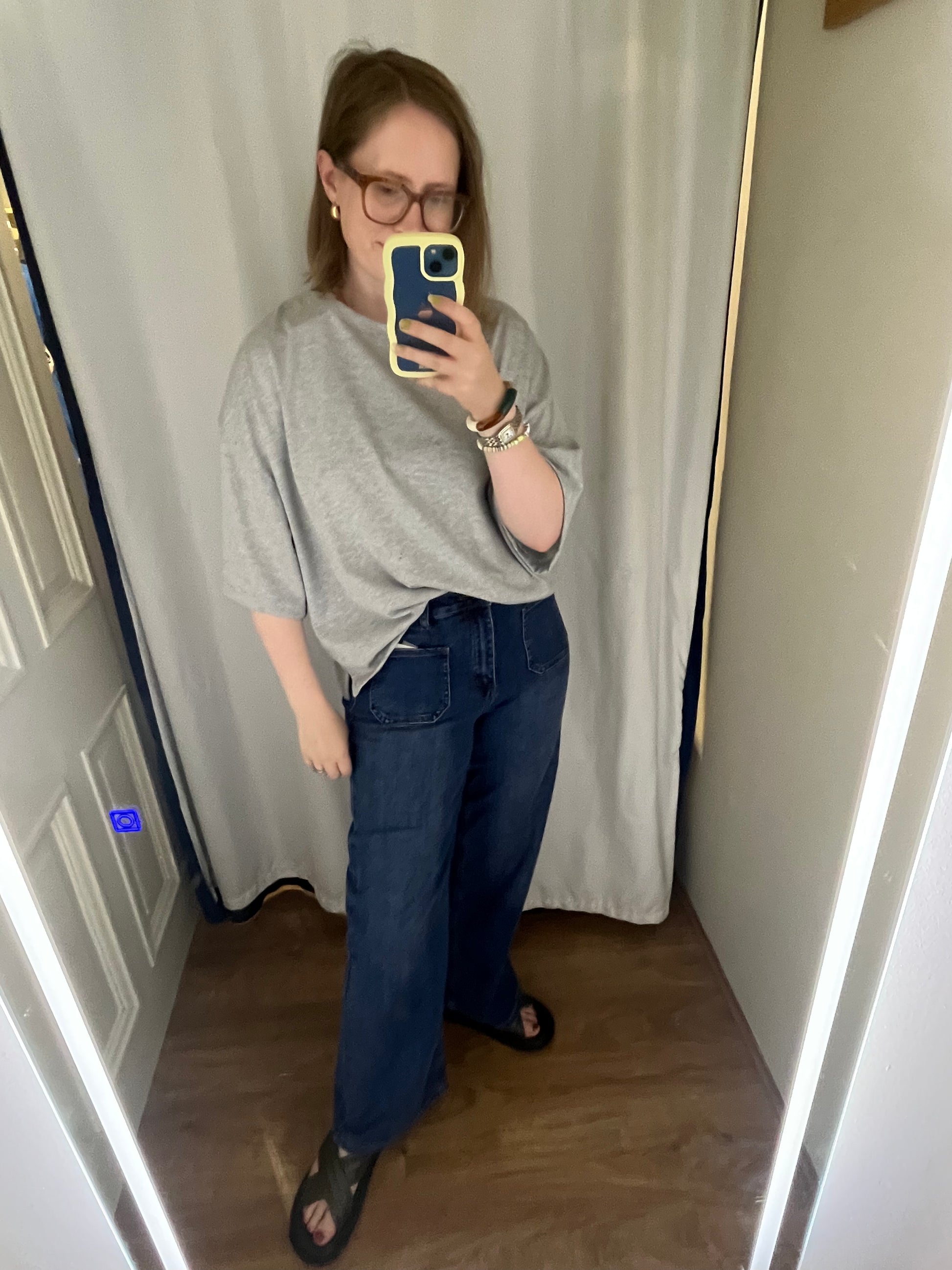 Person taking a mirror selfie wearing a gray sweater and blue jeans indoors.
