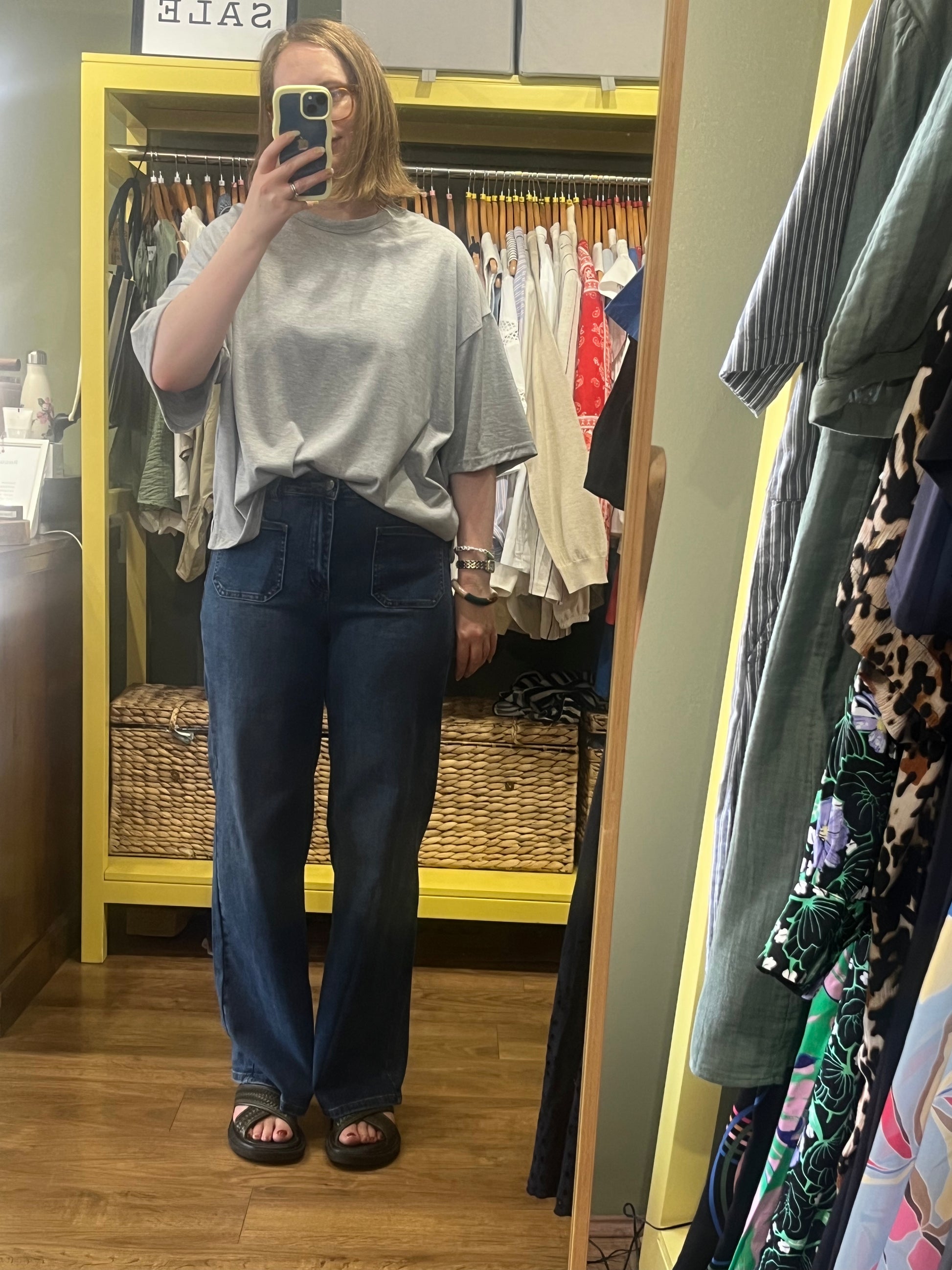Person taking a mirror selfie wearing a gray top and blue jeans in a clothing store.