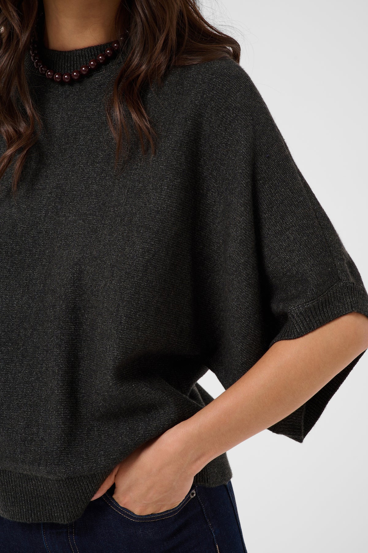 A close-up of a woman wearing a dark charcoal gray knit sweater with wide, short sleeves and ribbed trim, paired with dark blue jeans and a beaded necklace.