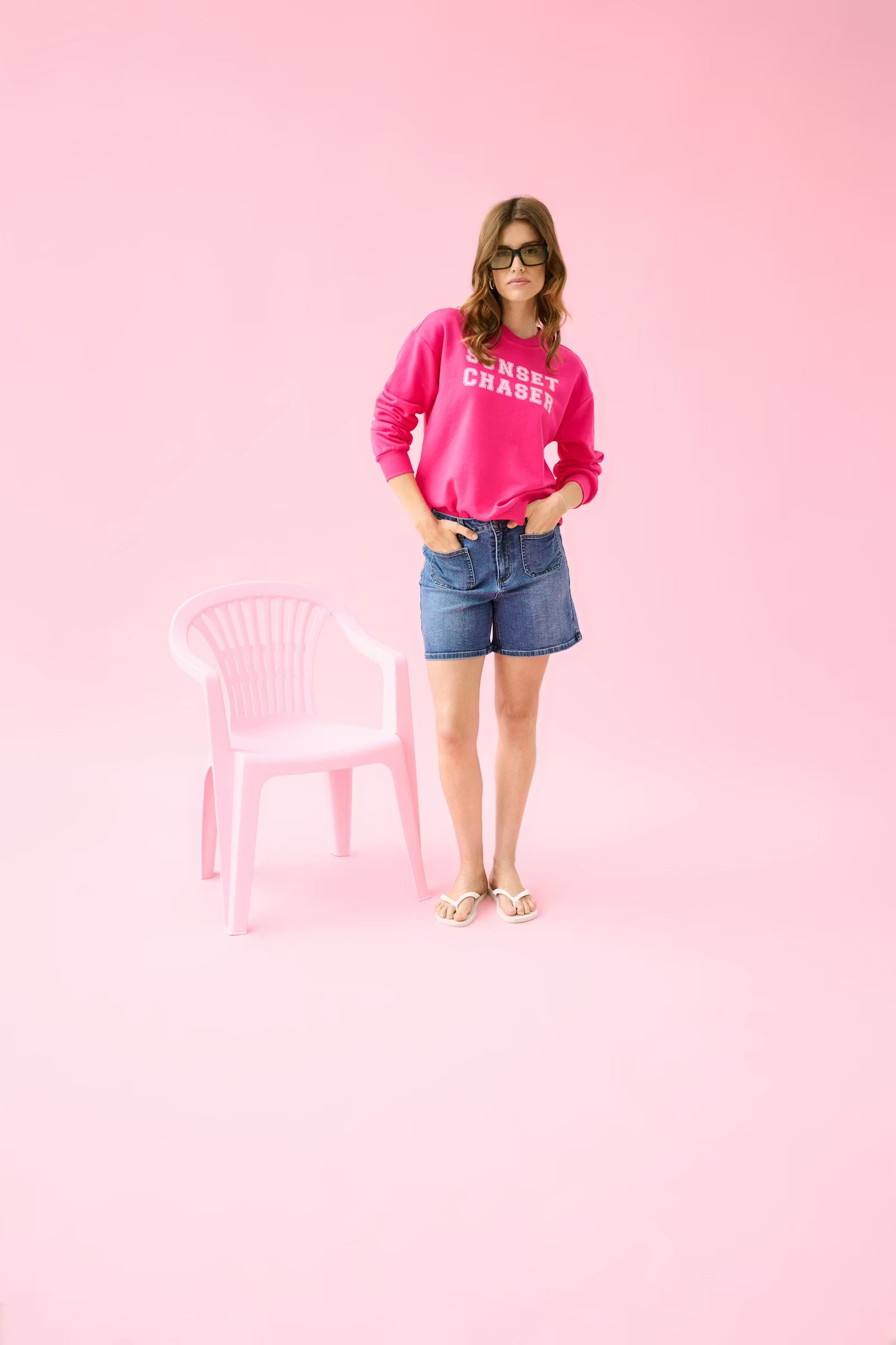 Woman wearing a pink sweatshirt and denim shorts standing against a pink background with a pink chair.