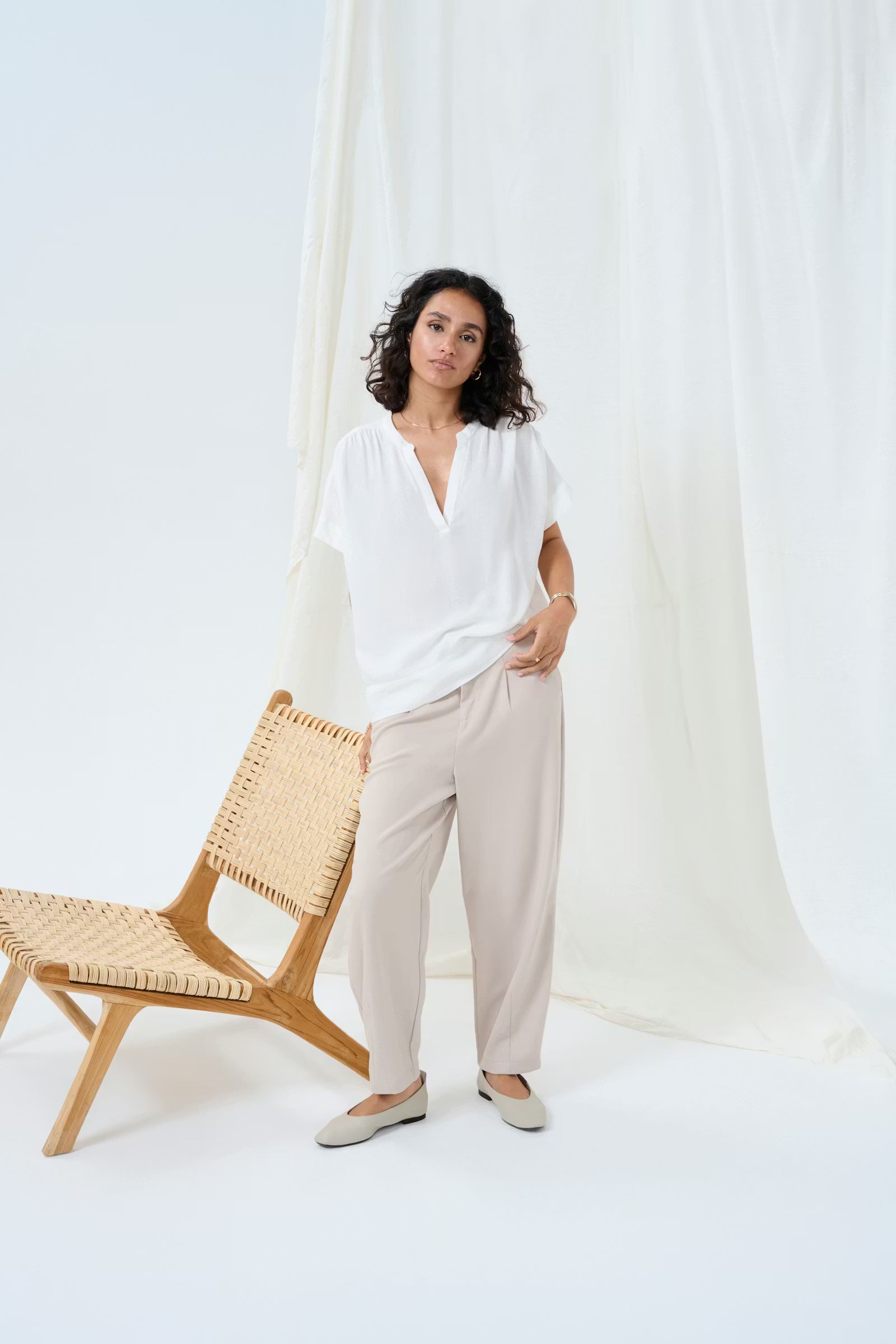 Woman wearing a white top and beige pants standing next to a wooden chair against a white curtain.