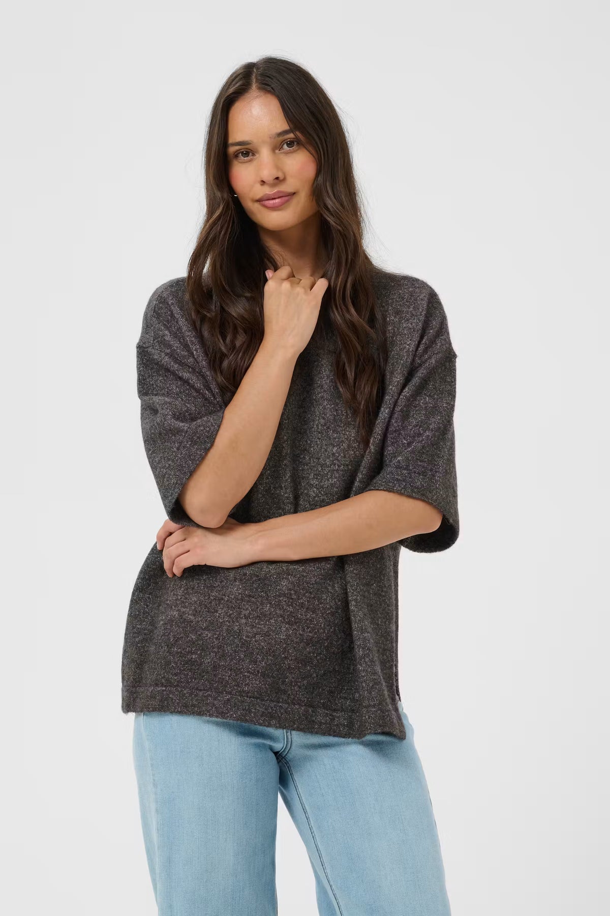 A woman modeling a dark gray heathered knit poncho-style top with light blue jeans against a plain light gray studio background.