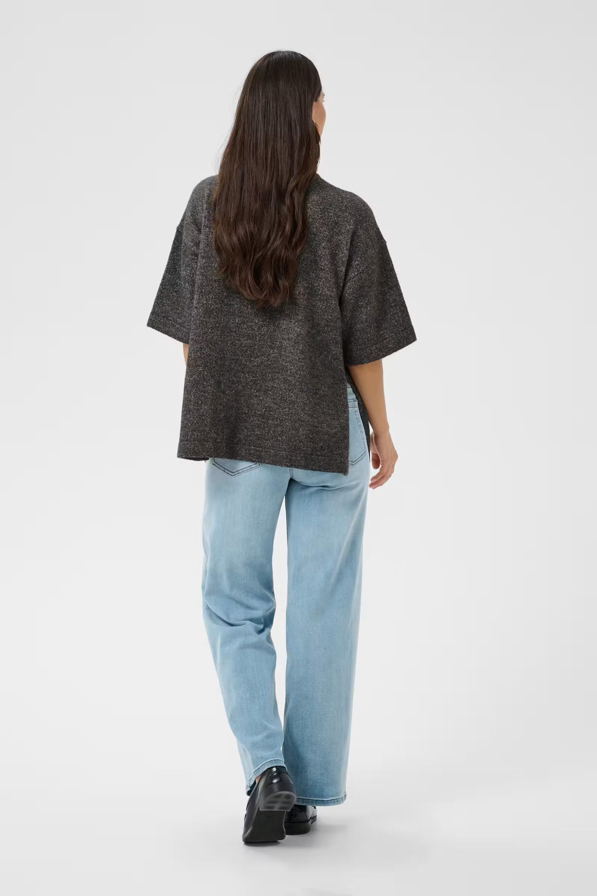 A woman shown from behind wearing a dark gray textured short-sleeve top, light blue wide-leg jeans, and black shoes, standing against a plain light gray studio background.