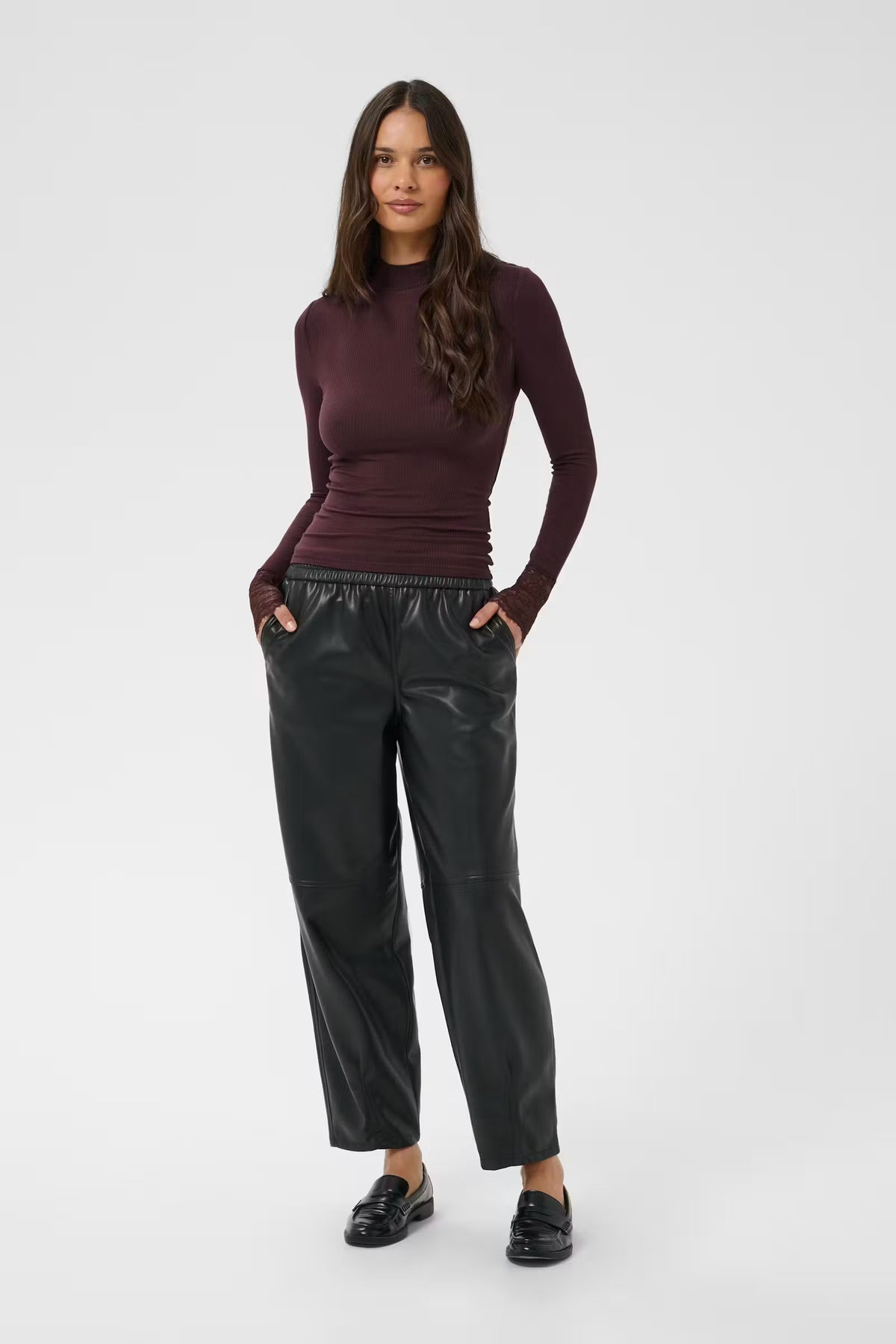 A woman modeling a dark plum long-sleeve mock neck top and black wide-leg faux leather pants against a light gray studio background.