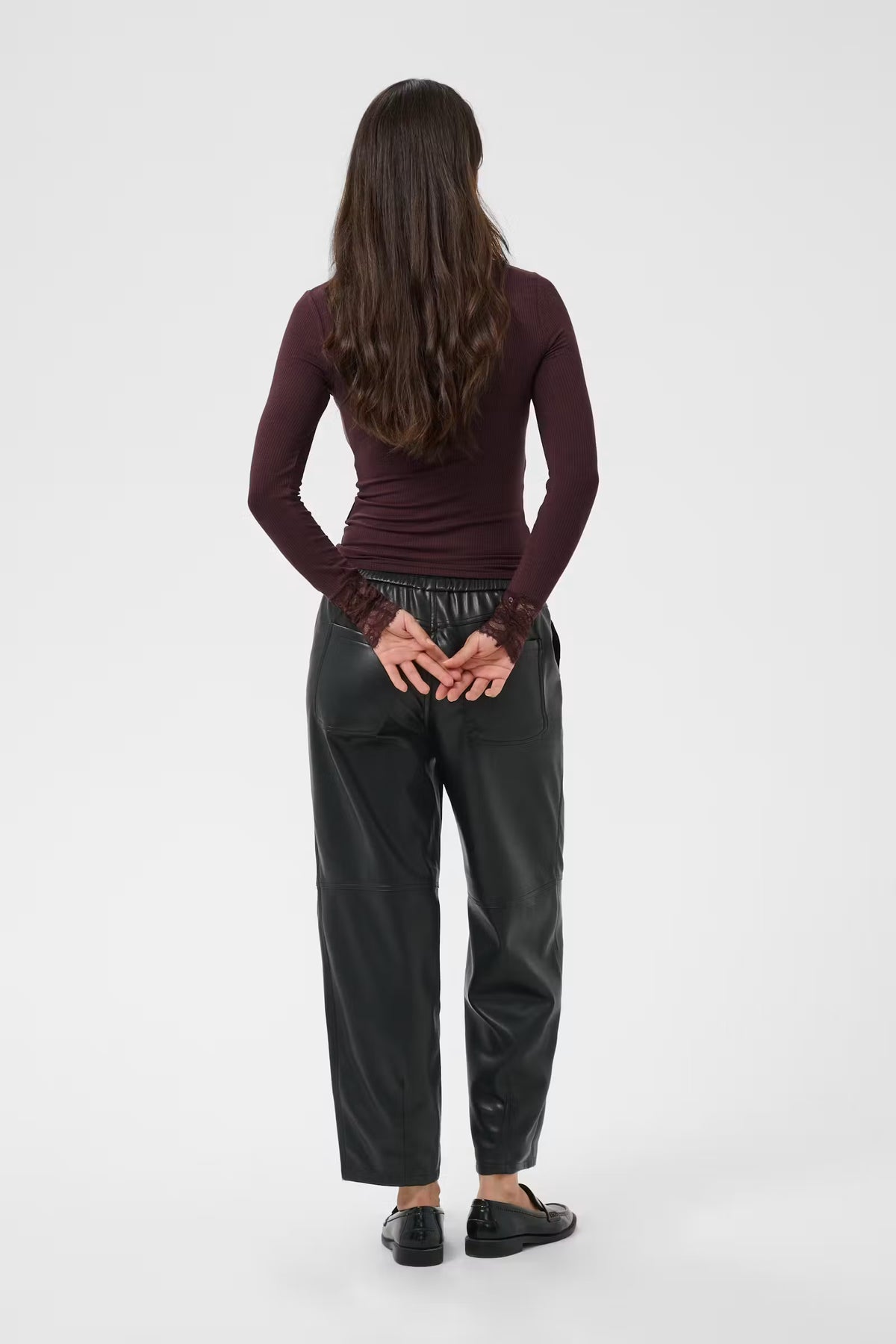 A woman viewed from behind wearing a dark burgundy long-sleeve top, black wide-leg pants, and black shoes against a light gray studio background.