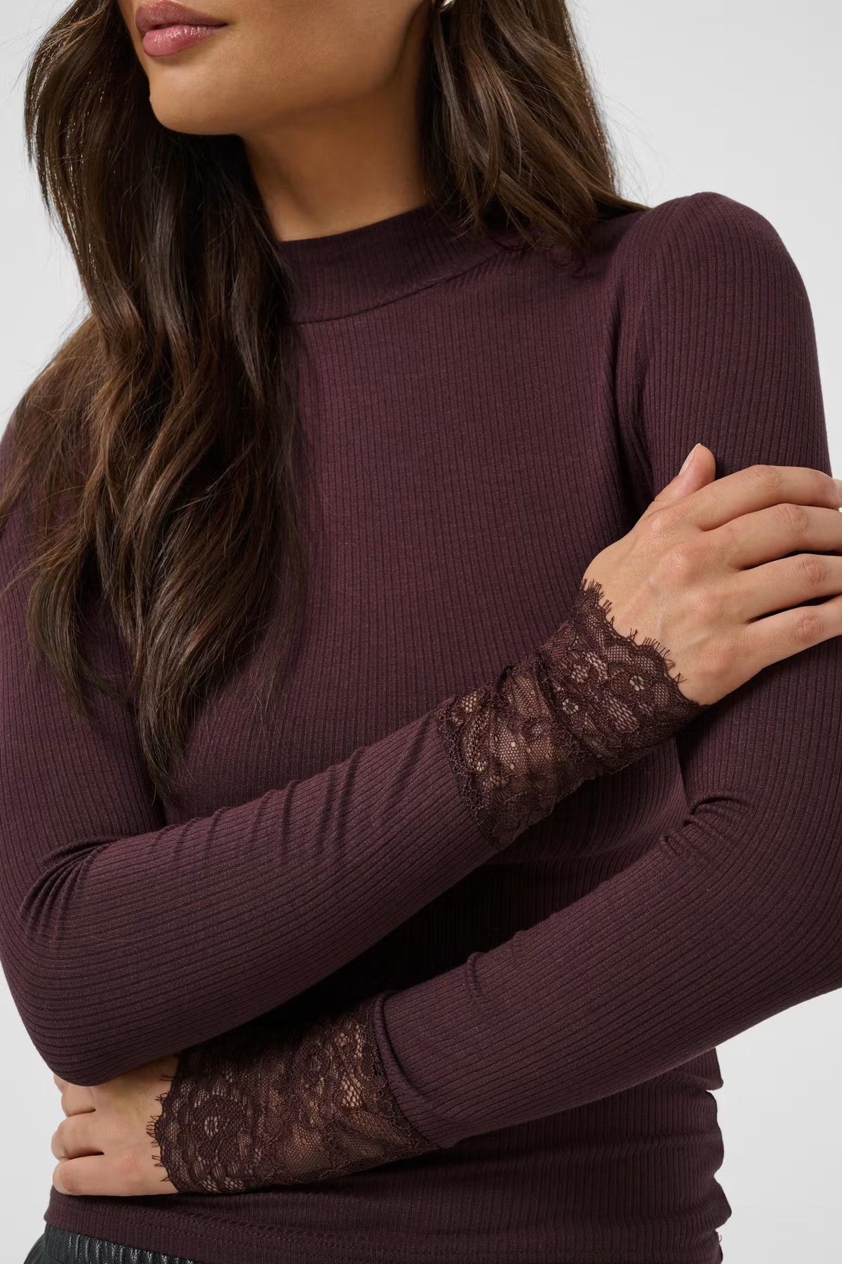 A close-up of a woman wearing a ribbed long-sleeve mock neck top in deep plum purple, featuring sheer lace cuffs on the sleeves.
