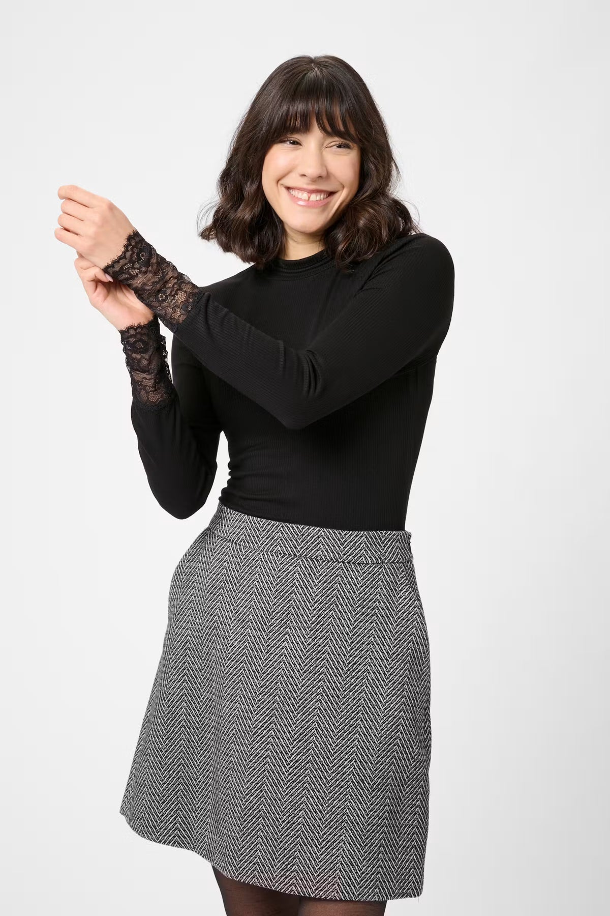 A woman modeling a black long-sleeve top with black lace cuffs and a gray and black herringbone skirt, posing against a light gray studio background.