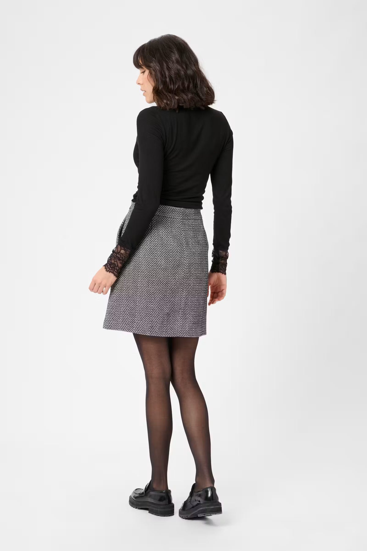 A woman is shown from the back wearing a black long-sleeve top with lace cuffs, a gray and white houndstooth mini skirt, sheer black tights, and black loafers, set against a plain light gray studio background.