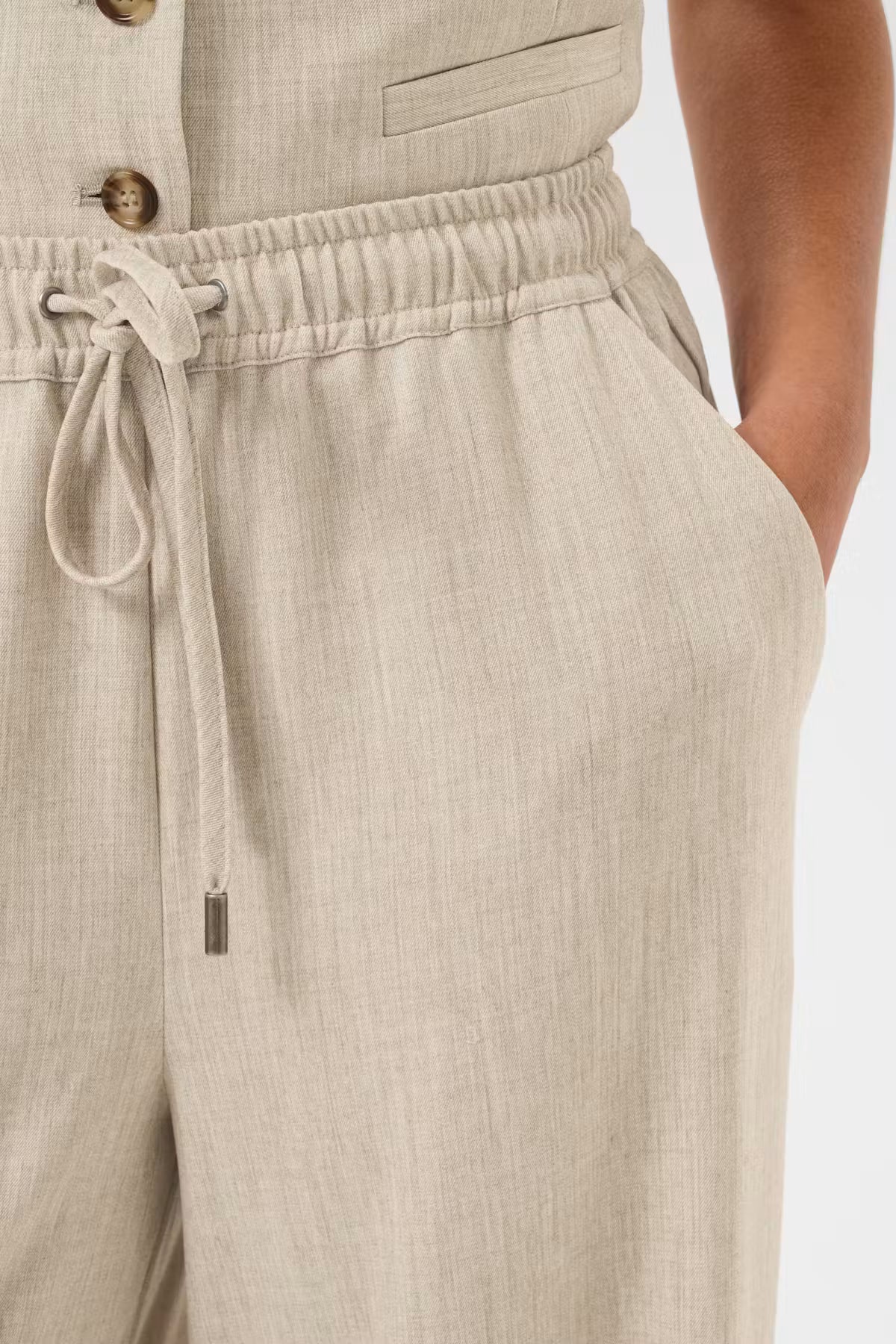 Close-up of a person wearing light beige linen drawstring trousers, showing the waistband, drawstring, side pocket, and part of a matching top.