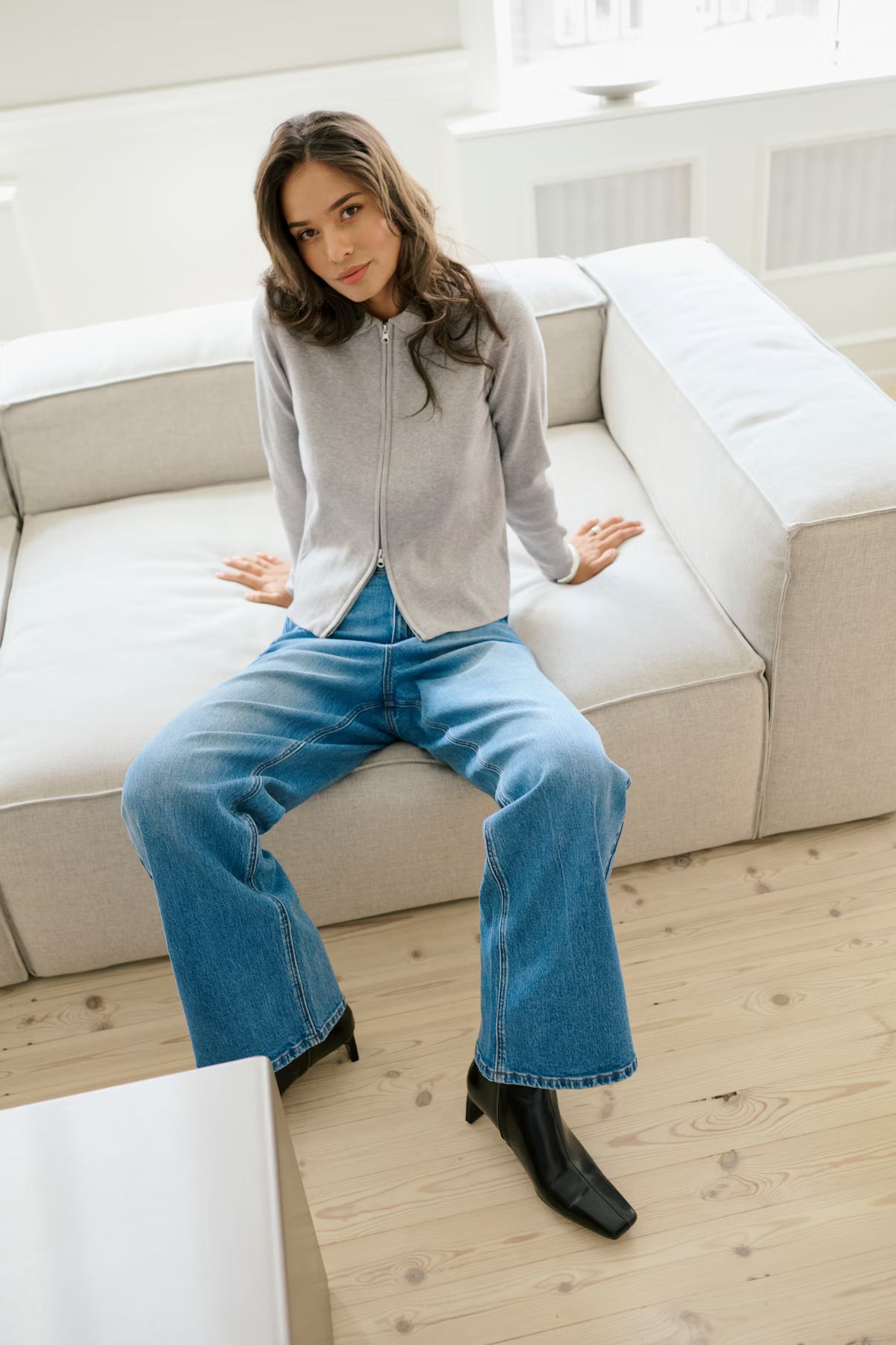 Woman sitting on a beige sofa wearing a gray zip-up hoodie, blue jeans, and black boots in a bright room.