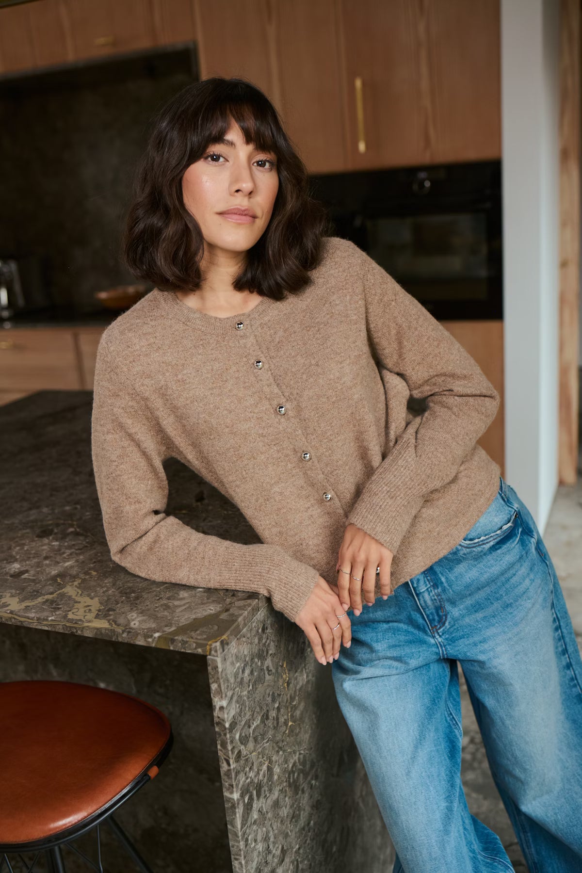 Woman in a beige cardigan and blue jeans leaning against a kitchen counter.