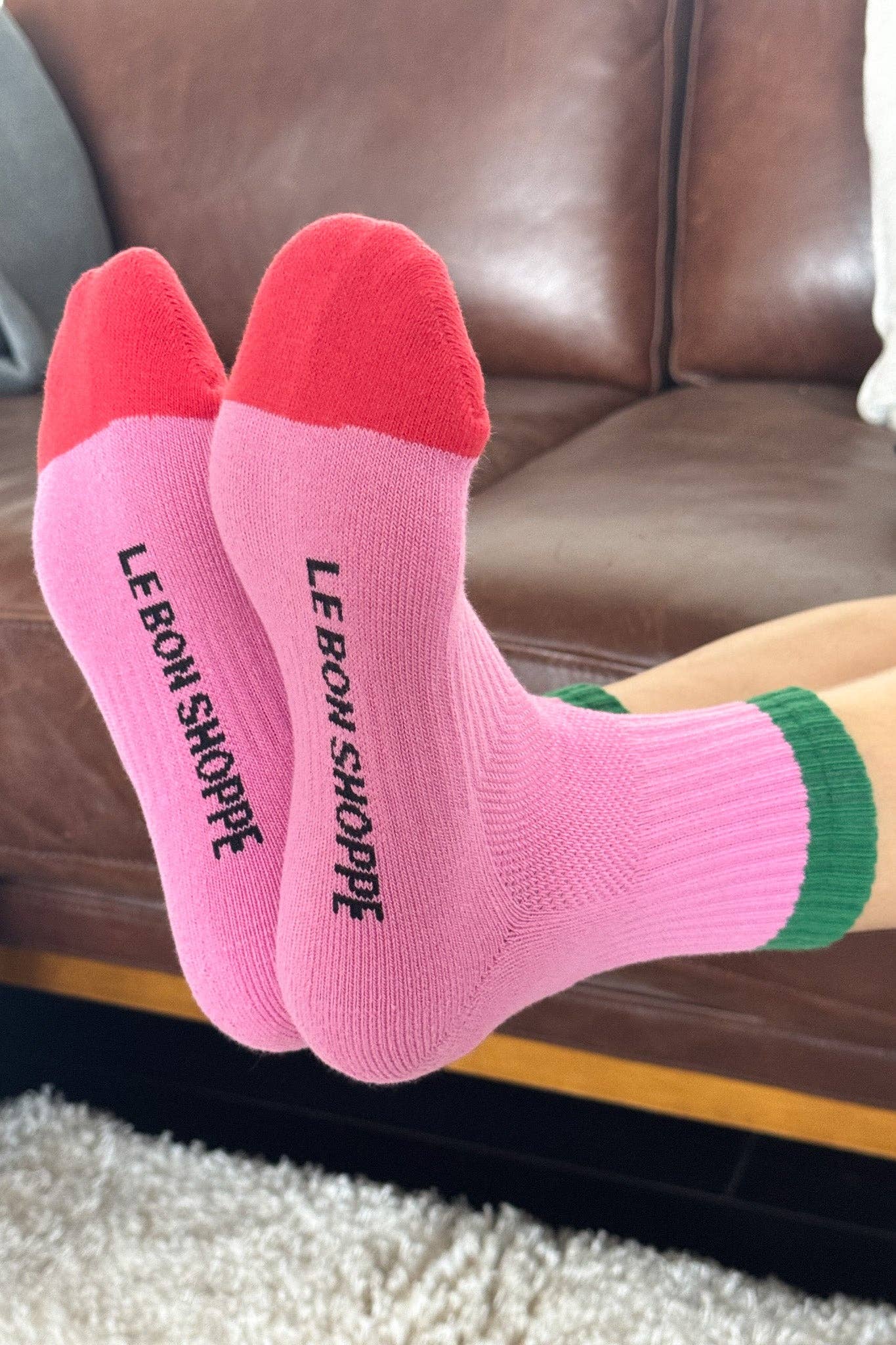 Pink socks with 'Le Bon Shoppe' text on a person sitting on a brown couch.