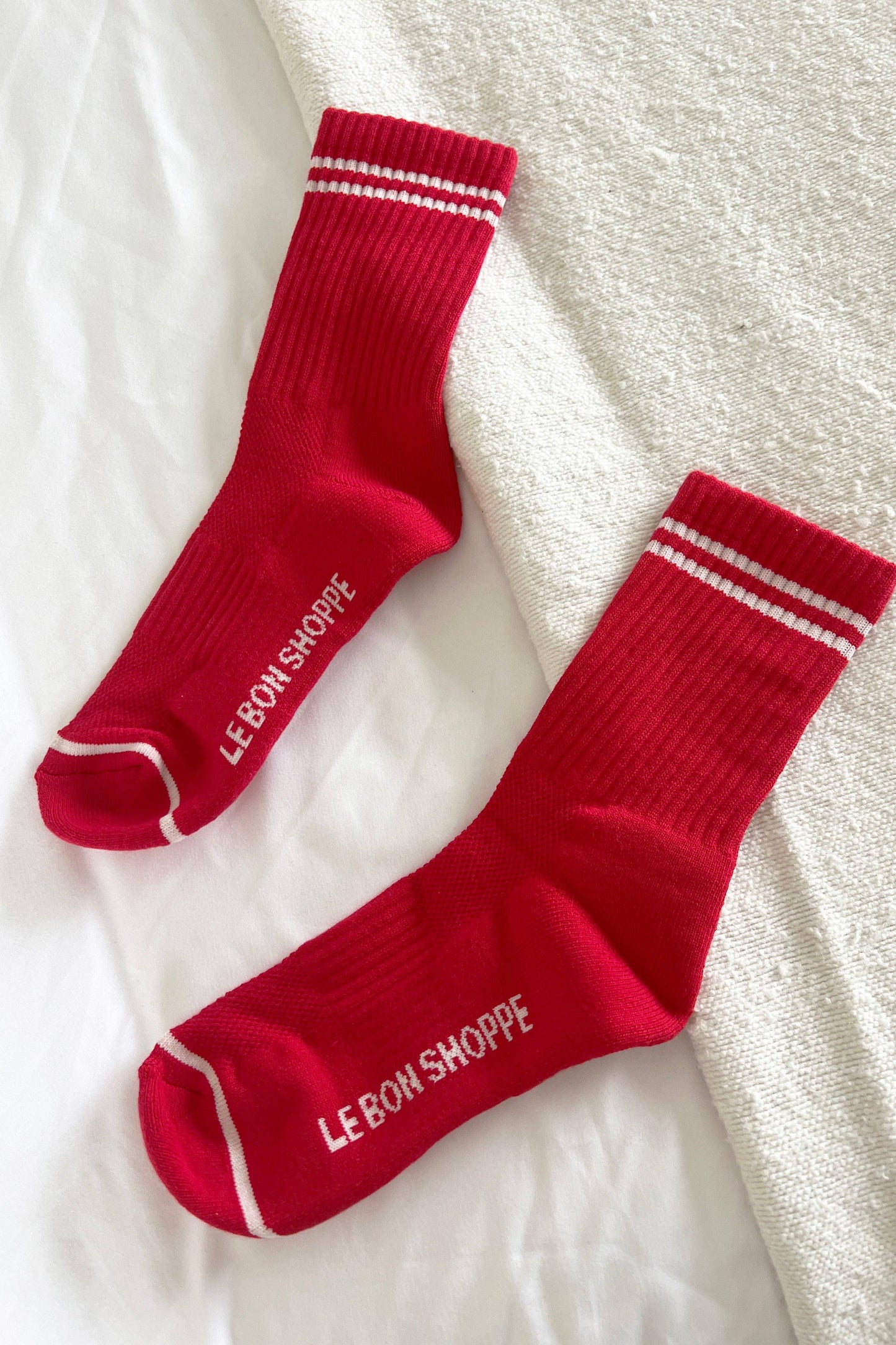 Two red socks with white stripes and 'Le Bon Shoppe' branding on a textured white surface.