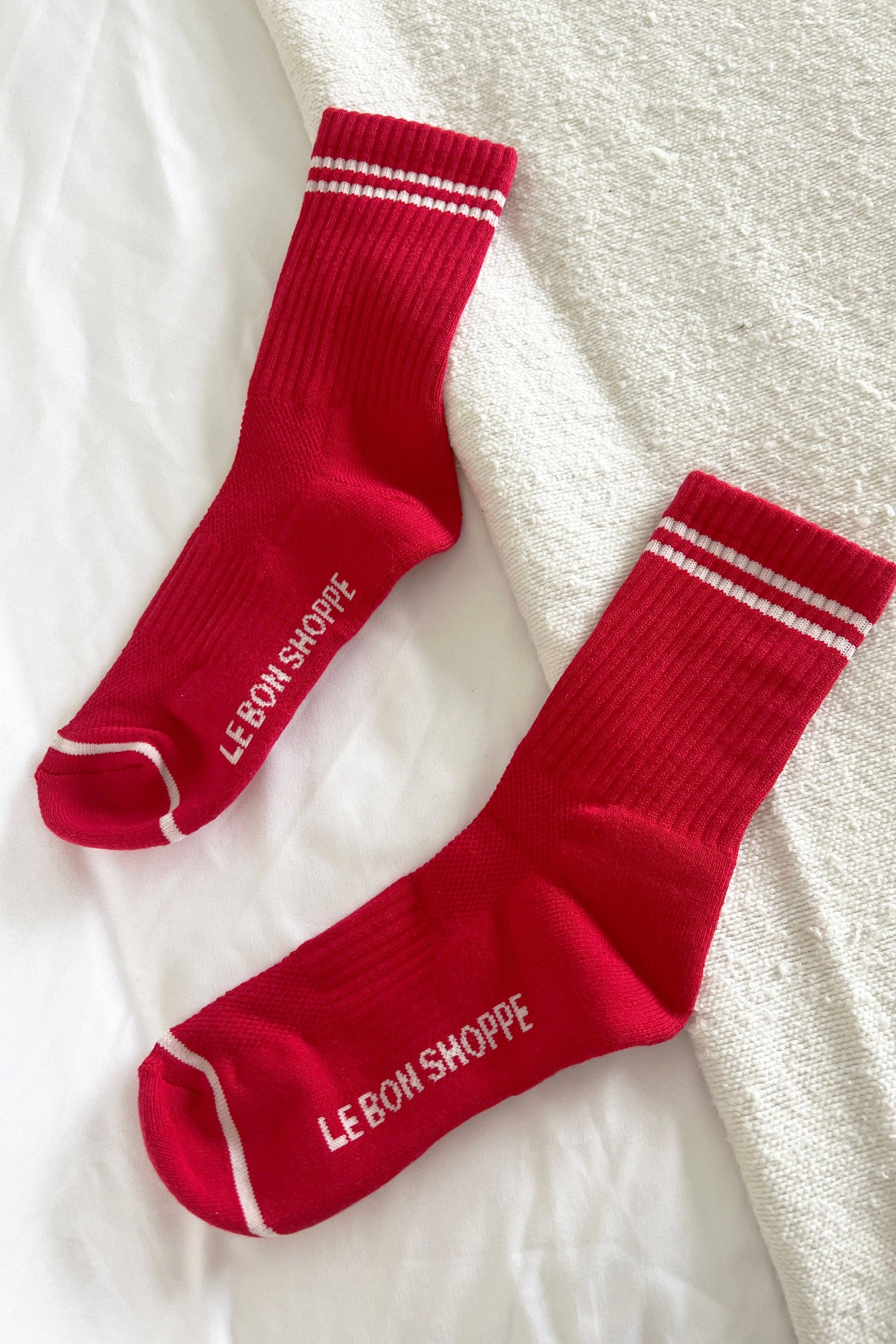 Two red socks with white stripes and 'Le Bon Shoppe' branding on a textured white surface.
