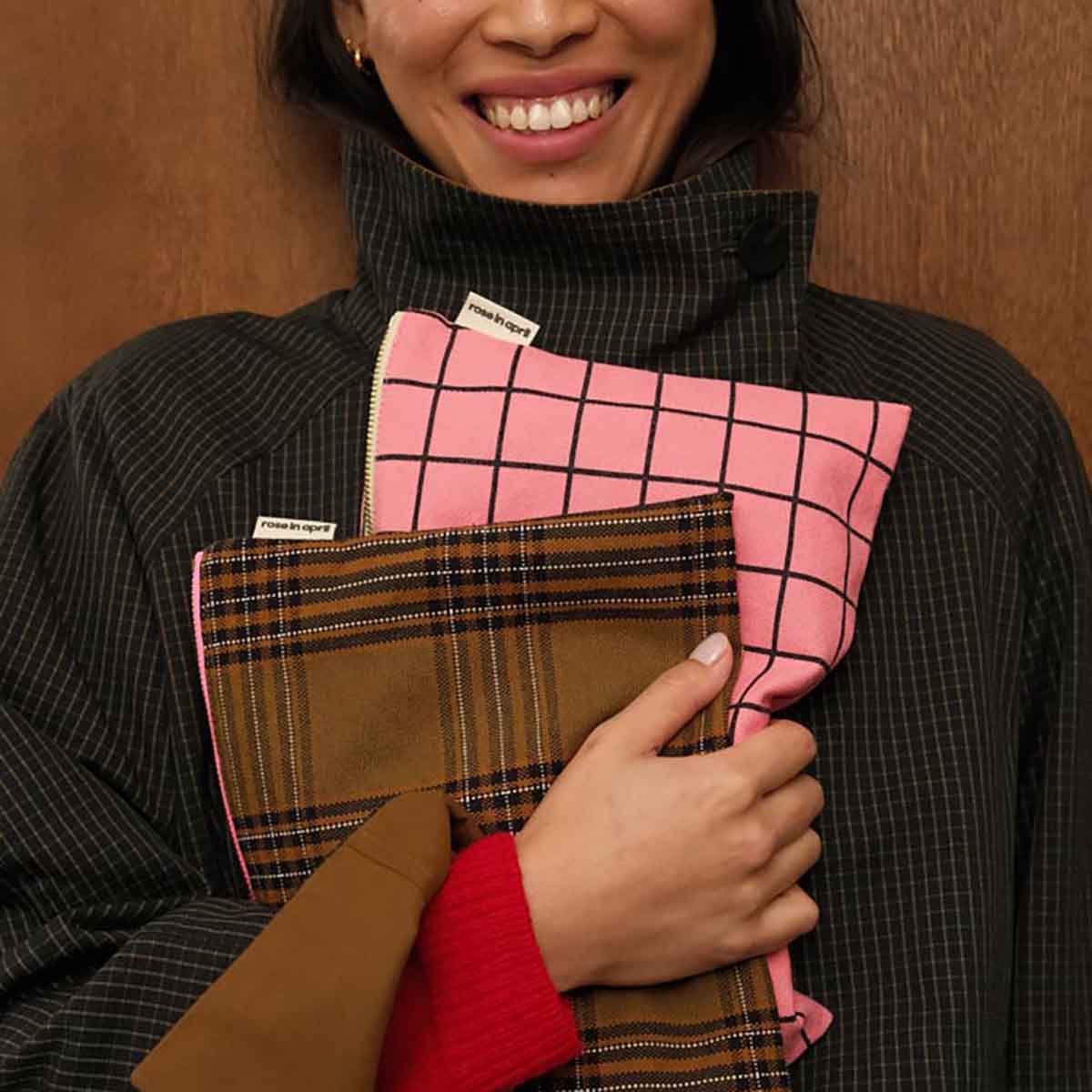 Person holding two patterned bags, one pink with black grid and one brown plaid, against a wooden background.