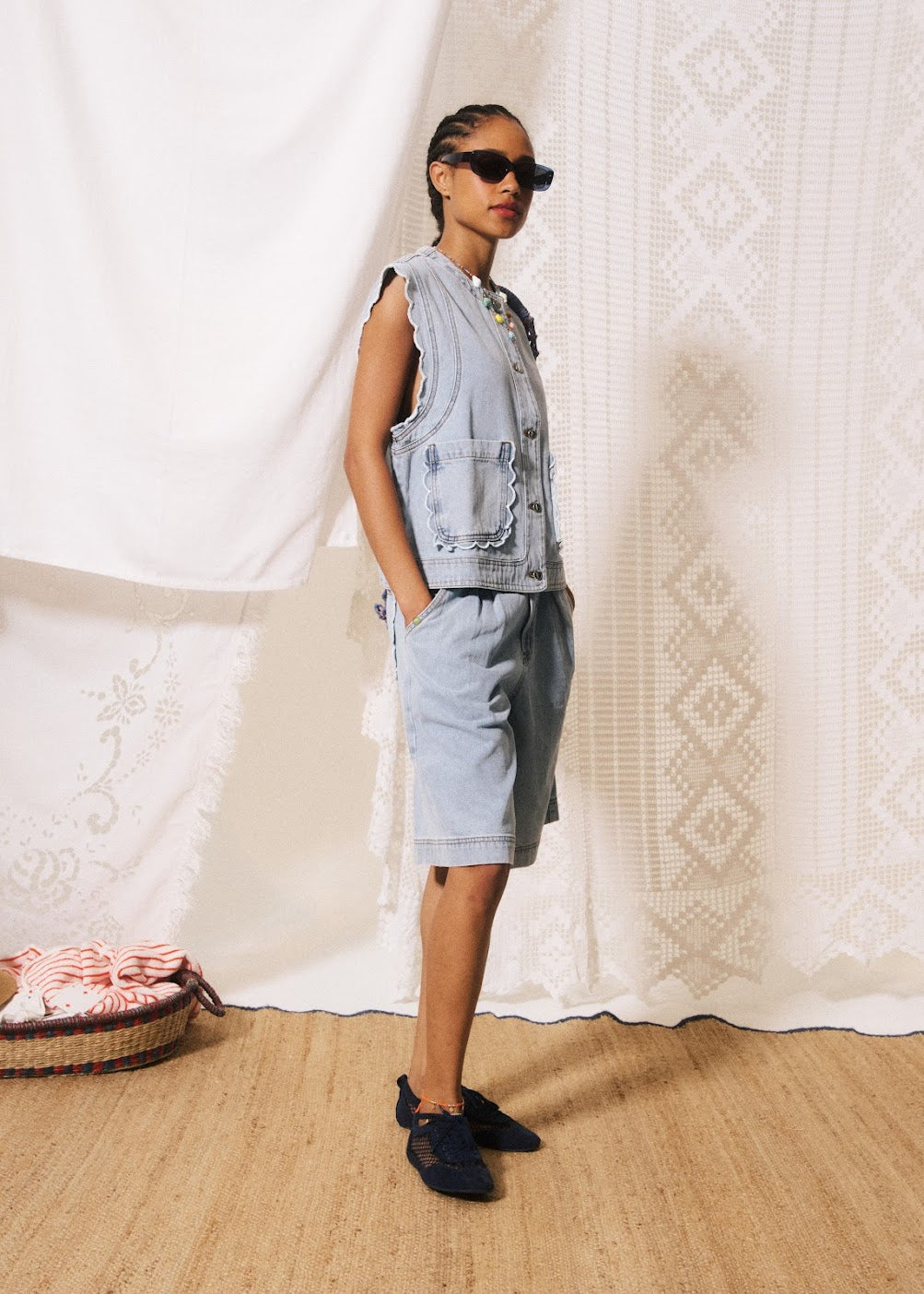 Woman wearing a denim vest and shorts standing in front of a textured white wall.