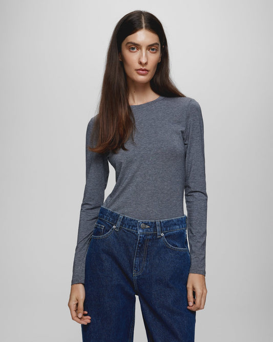 A woman modeling a dark gray long-sleeve crewneck top paired with dark blue jeans, photographed against a neutral light gray studio background.