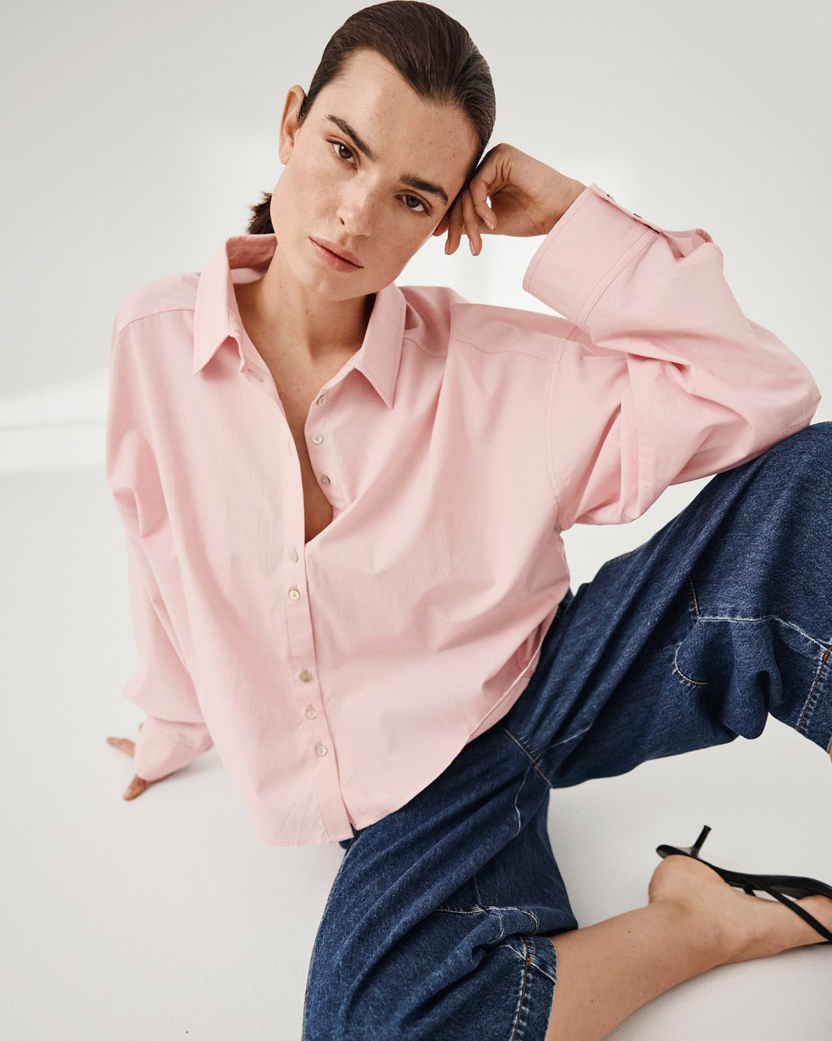 Woman wearing a pink shirt and blue jeans sitting on a white surface.