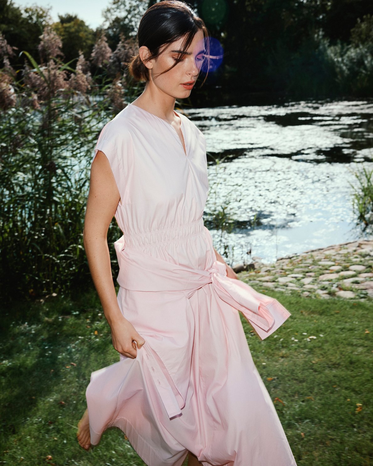 Woman in a light pink dress standing by a body of water with greenery.