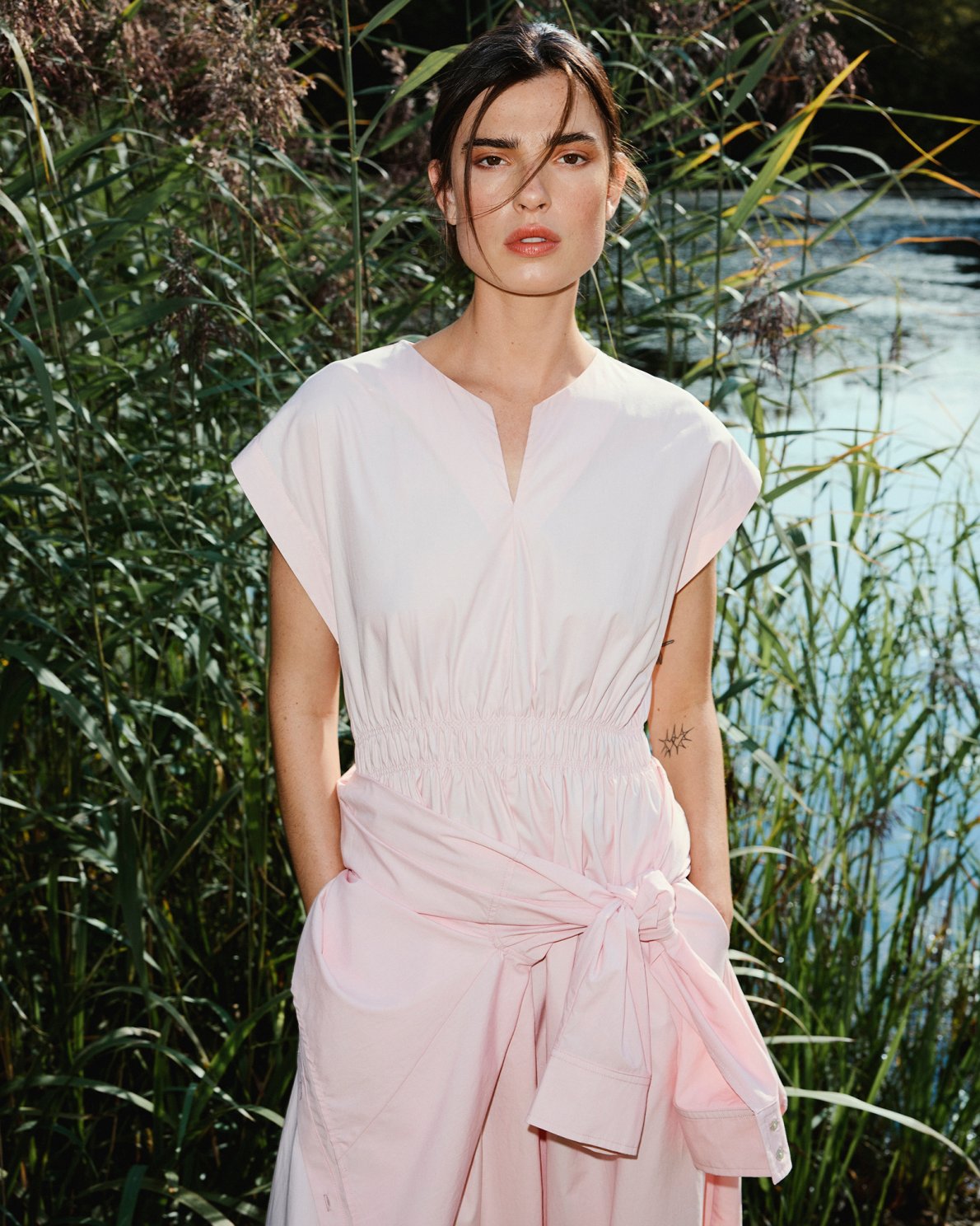 Woman in a light pink dress standing in a natural setting with water and greenery.