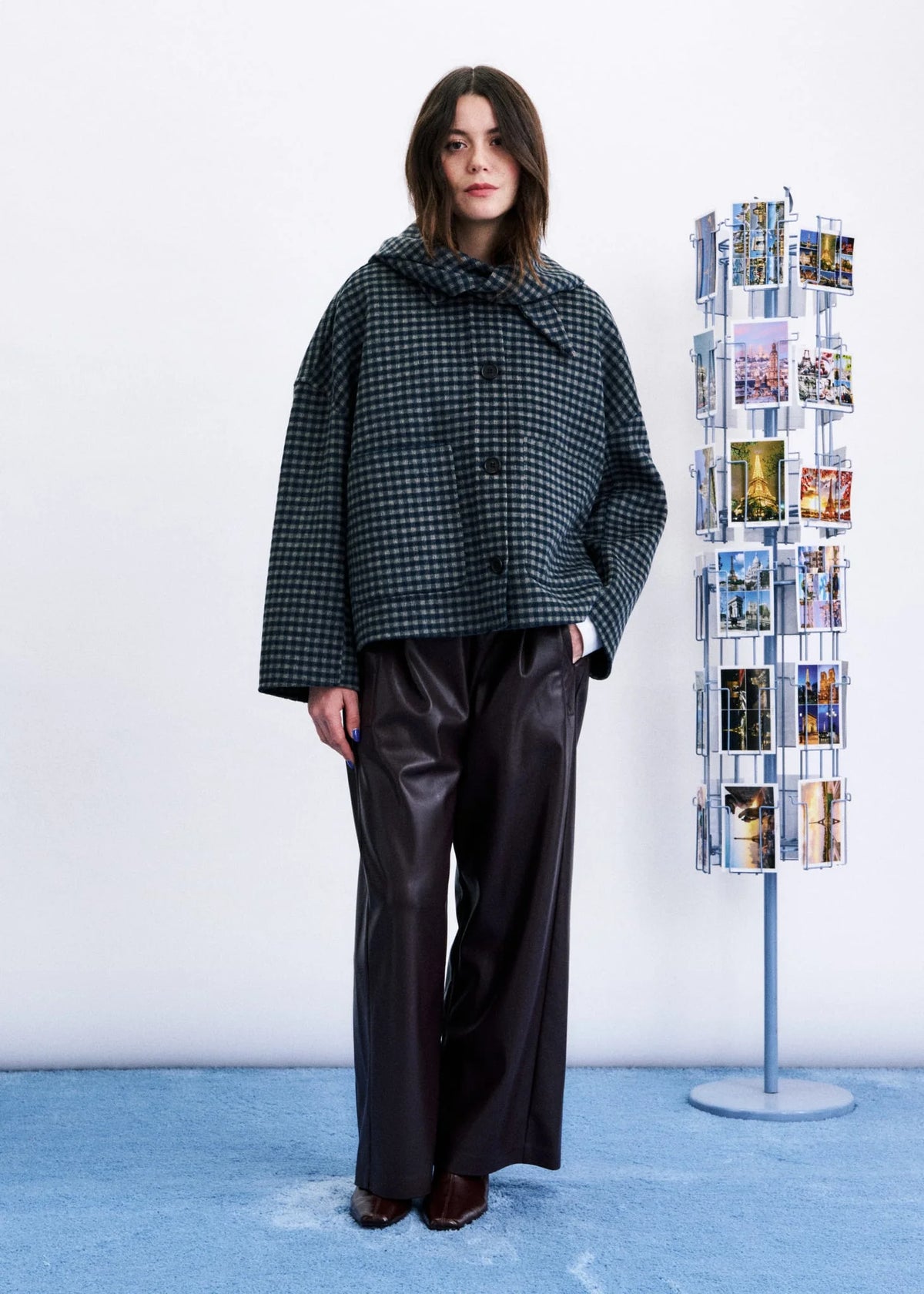 A model stands in a studio wearing a dark green and black checkered cropped jacket with a hood, paired with black wide-leg pants. To the right is a clear acrylic photo display stand against a white wall and light blue floor.